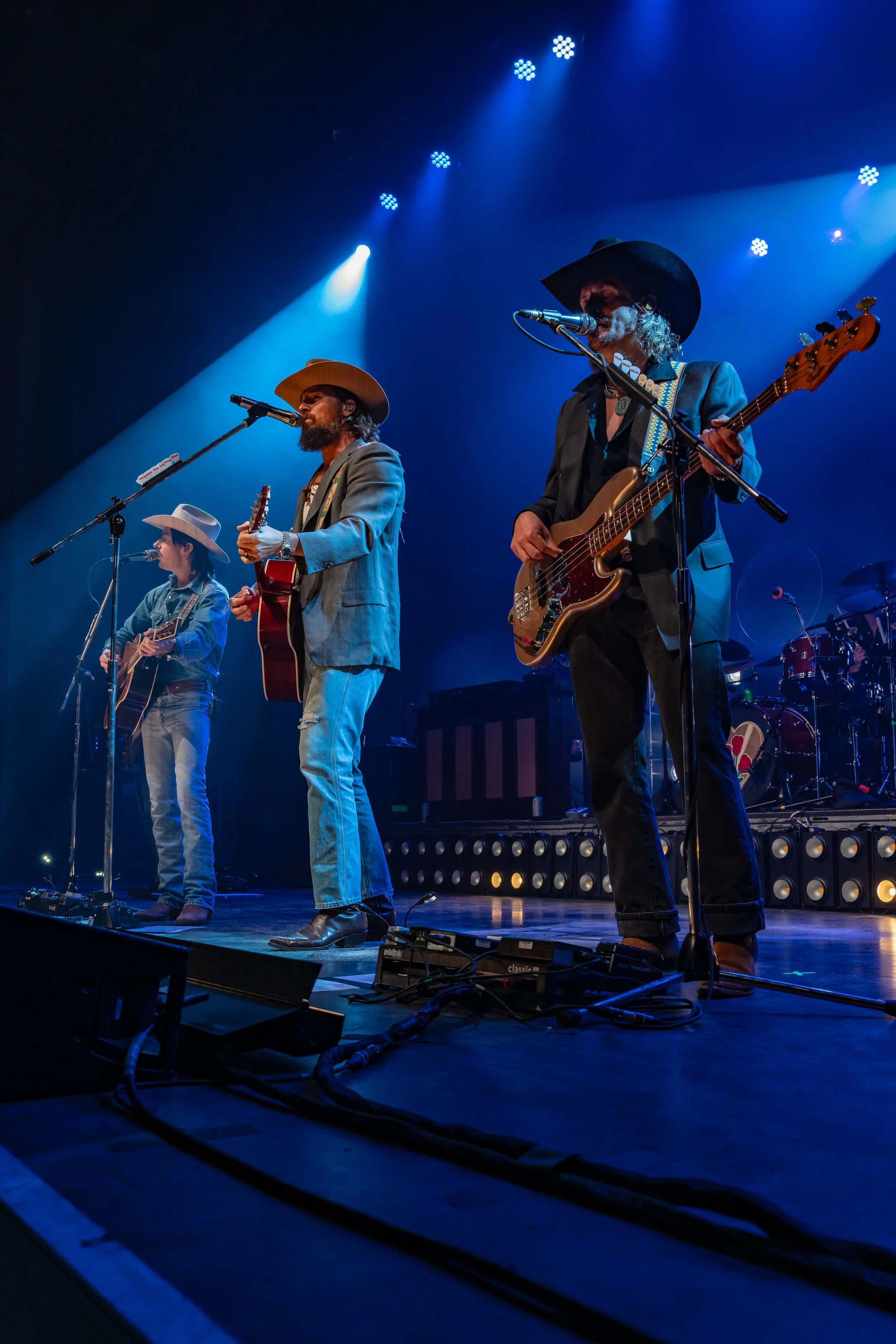 Three musicians performing on stage with guitars and microphones, under blue stage lights.