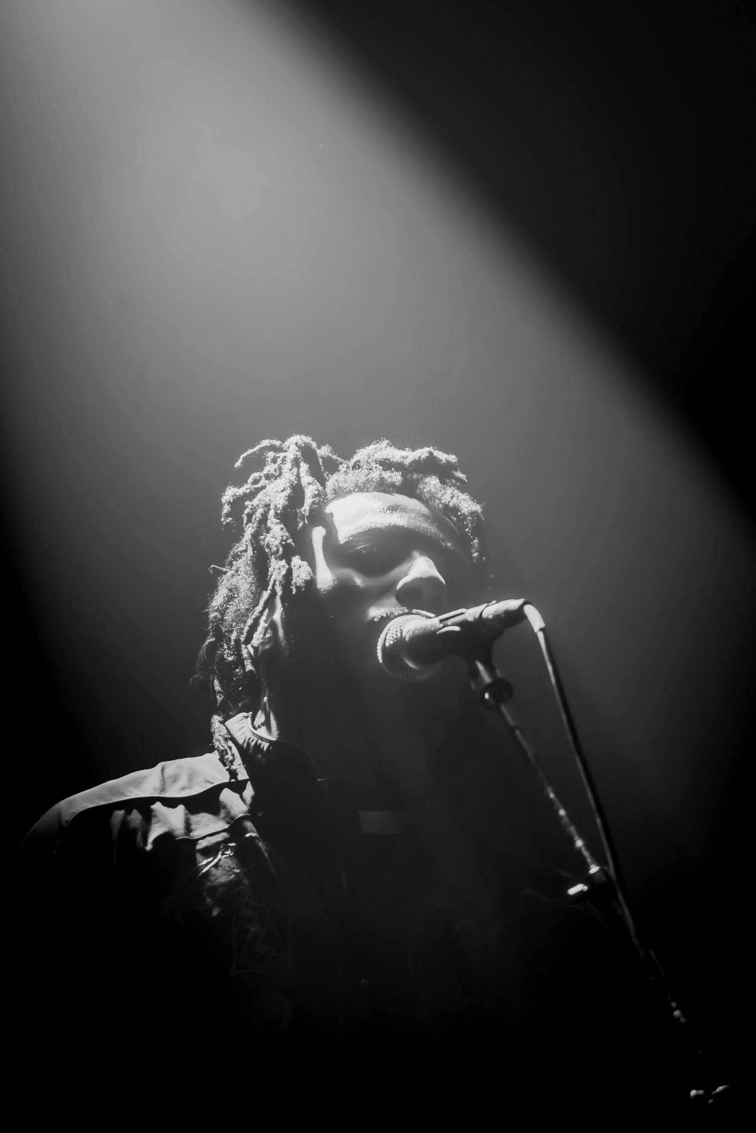 A black and white photo of a person with dreadlocks singing into a microphone on stage, illuminated by a spotlight.