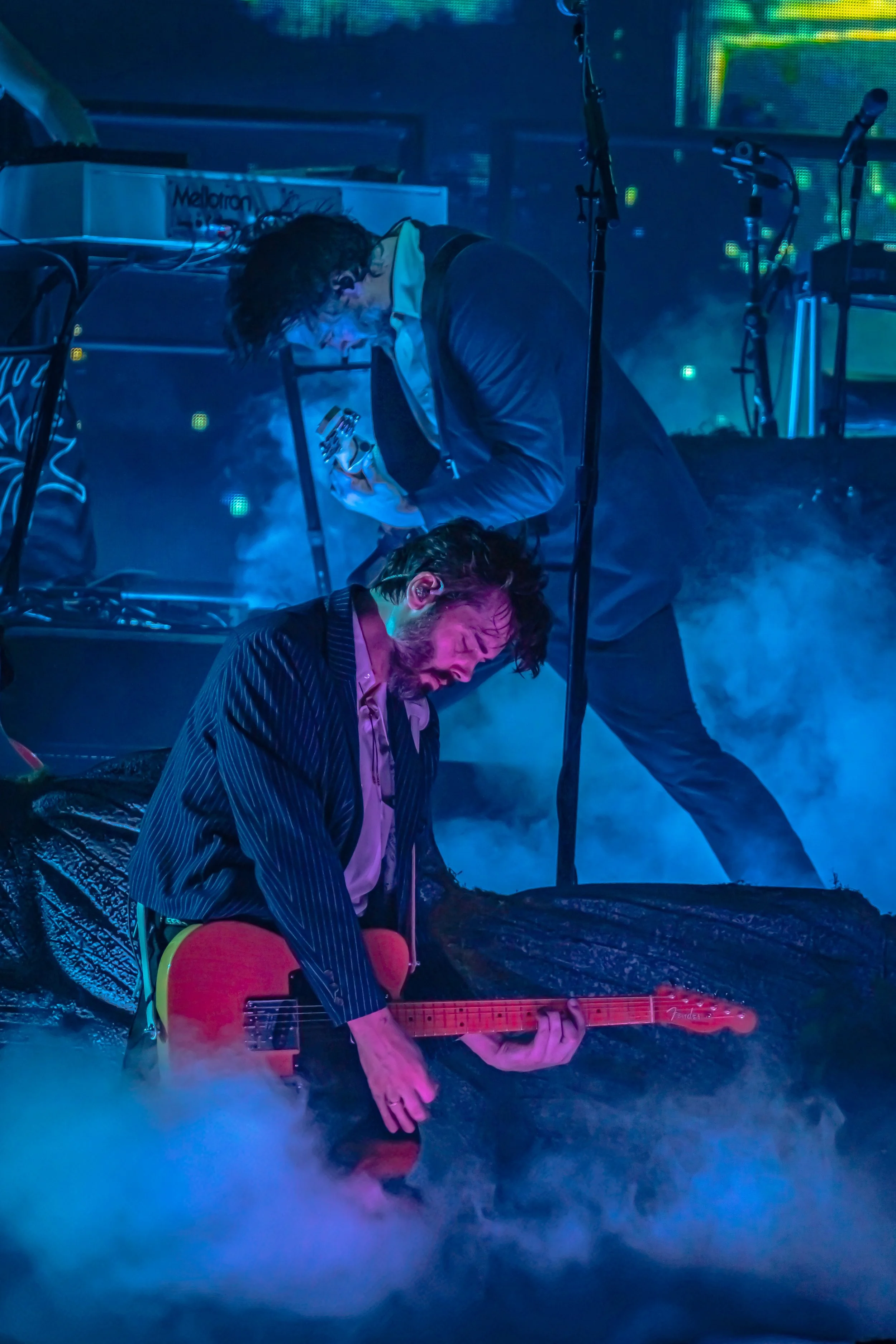 Two musicians performing on stage amid blue smoke; one is playing an electric guitar, kneeling, and the other is bent over a piece of equipment, possibly singing or adjusting settings, with microphones and equipment visible in the background.