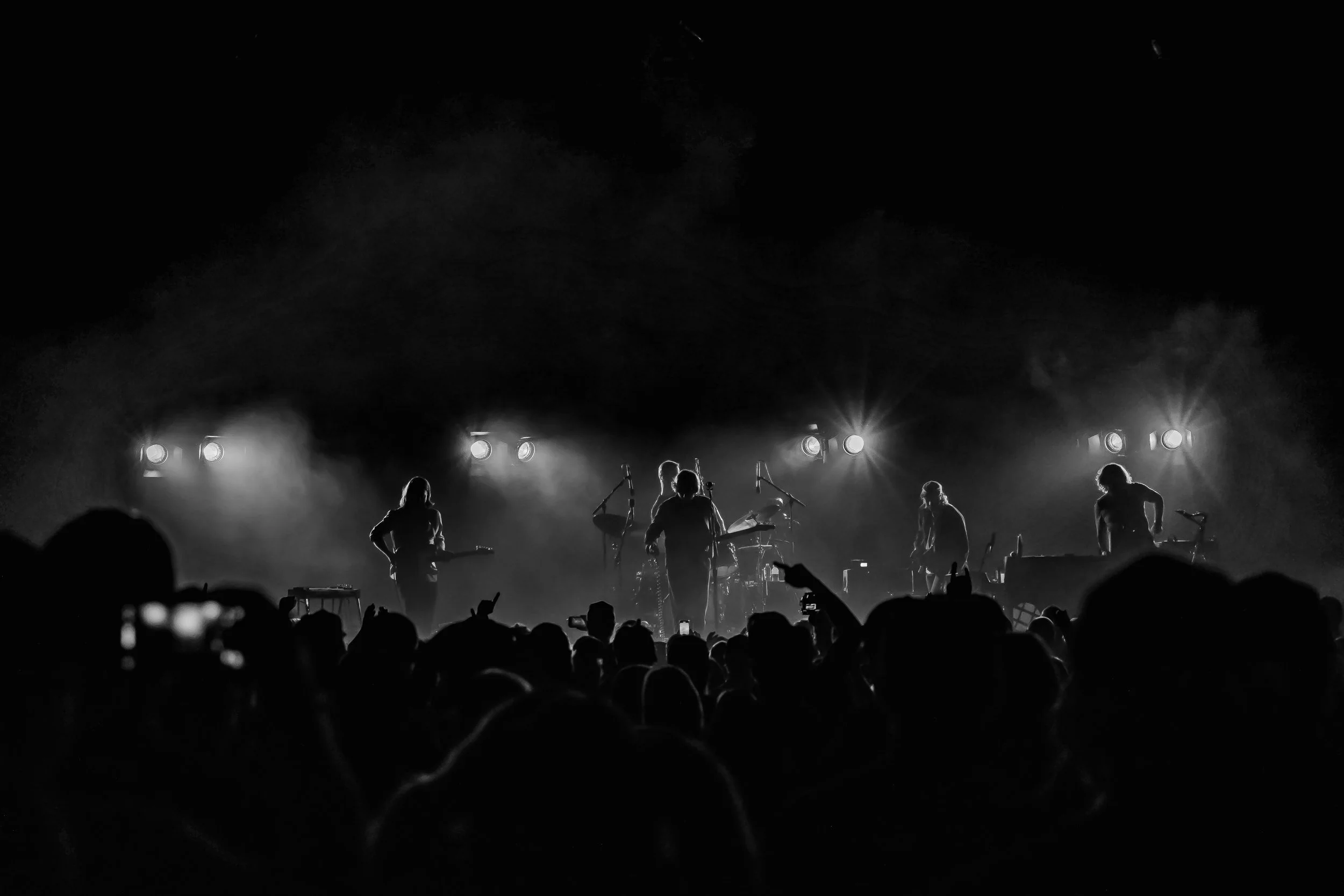 Black and white photo of a band performing on stage with bright lights shining behind them, audience silhouettes in front.