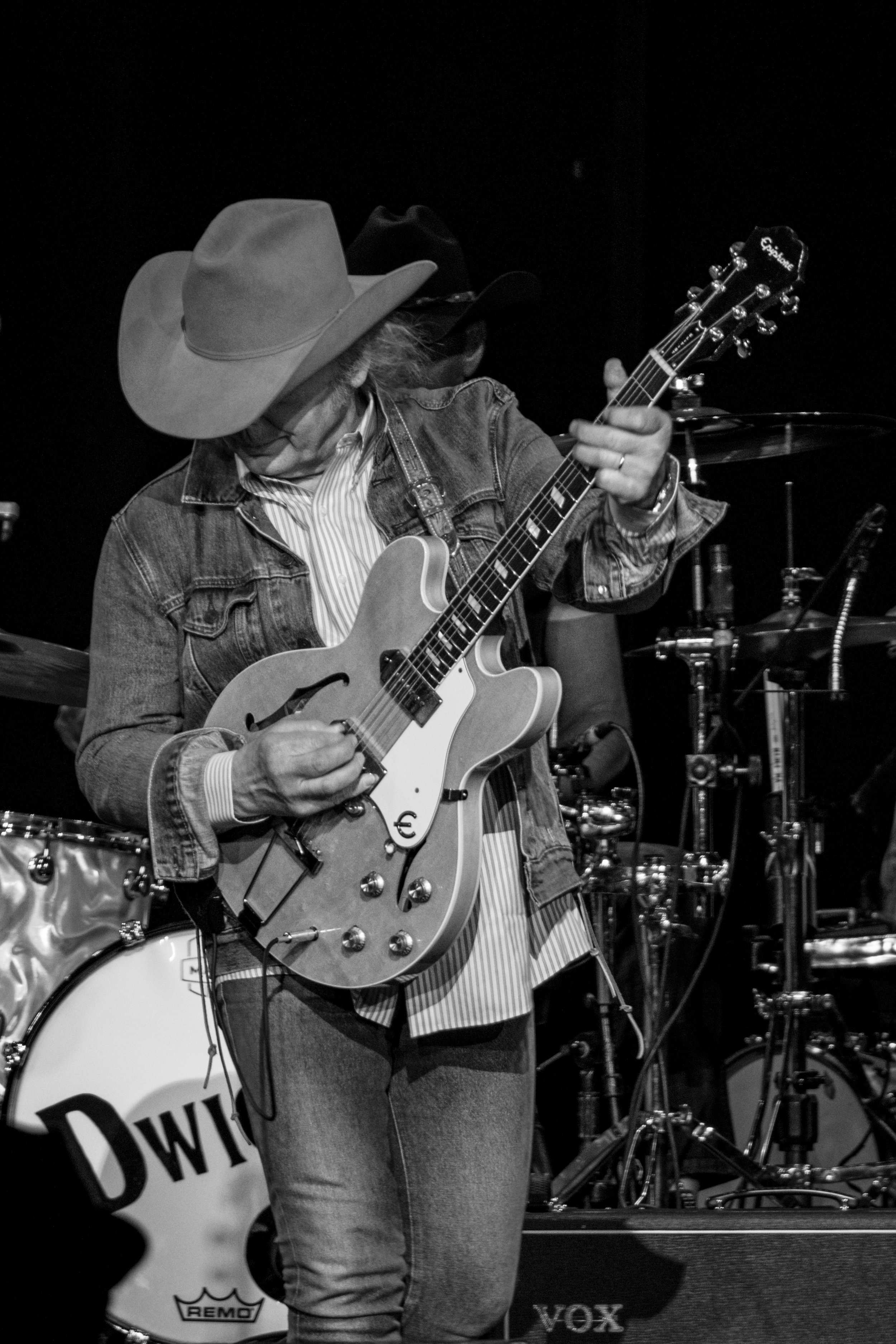 Black and white photo of a musician wearing cowboy hat, denim jacket, and striped shirt, playing an electric guitar on stage with drums in the background.
