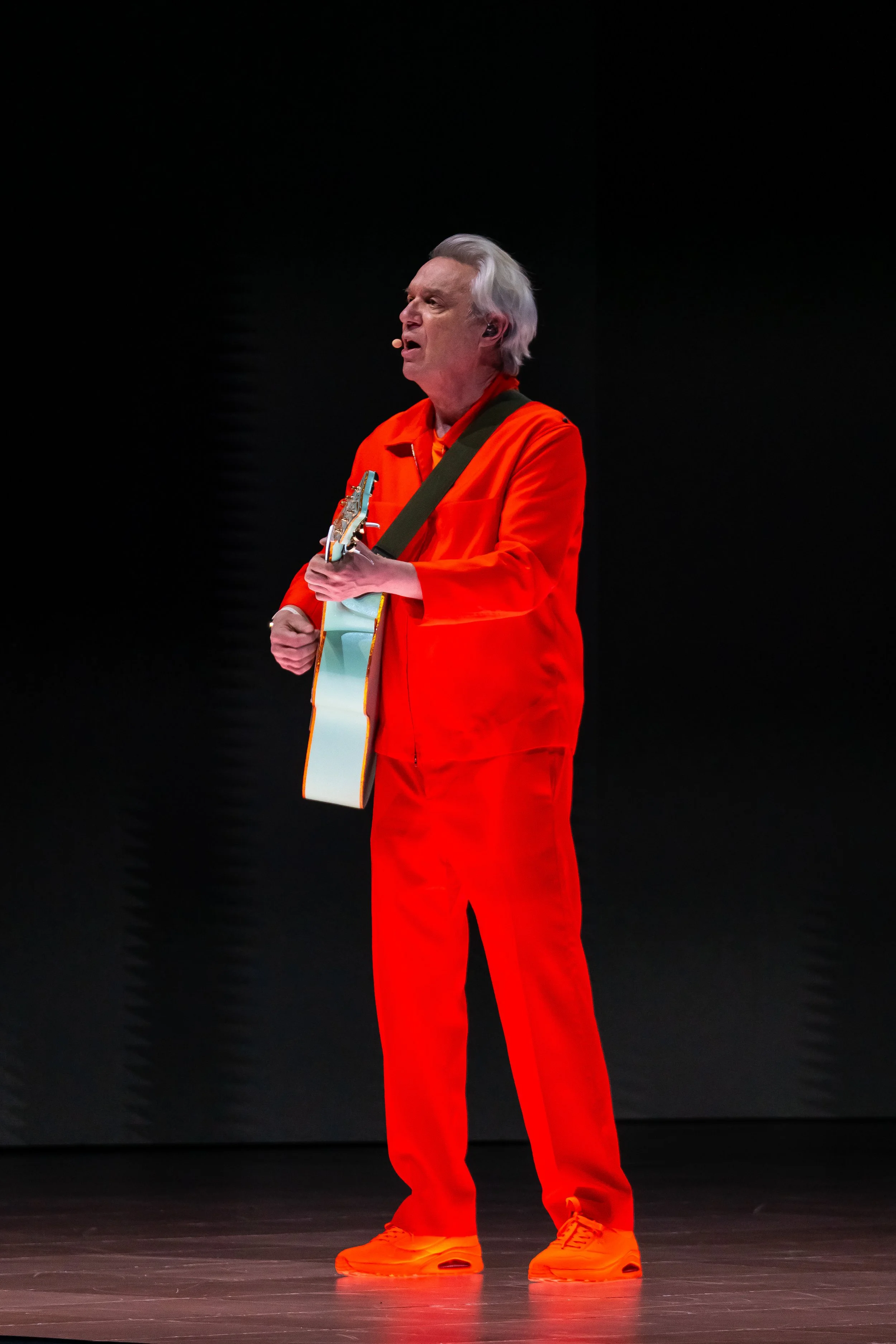 An older man with gray hair is standing on a stage, wearing an orange suit and orange sneakers, playing an electric guitar against a black background.