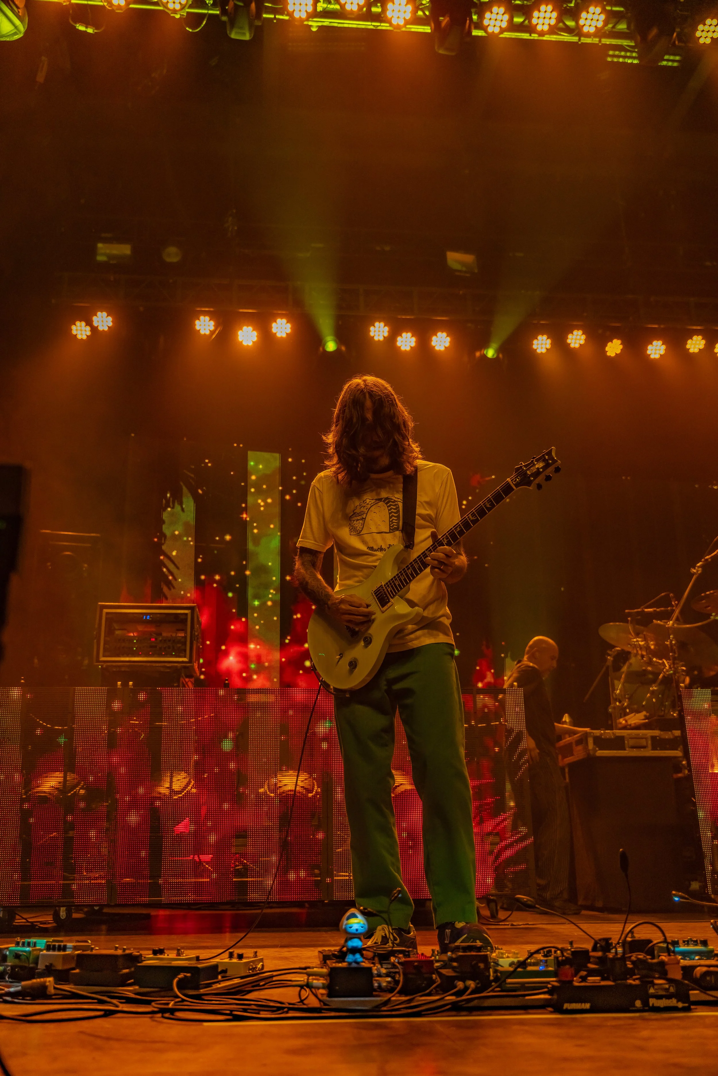 A musician with long hair playing an electric guitar on stage with colorful lights and digital displays behind him.