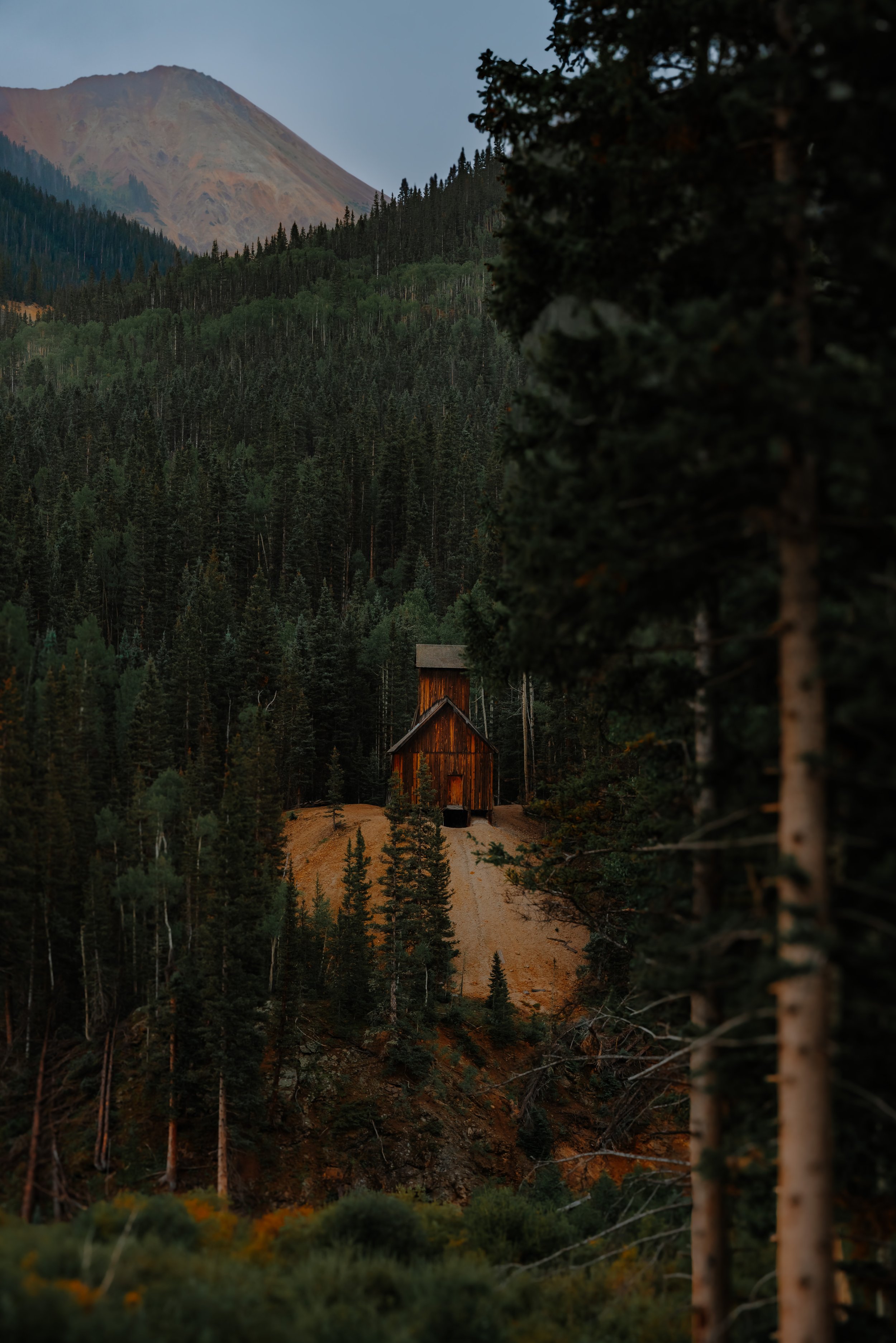 A wooden church surrounded by dense forest of tall pine trees, with mountain in the background during sunset or dusk.