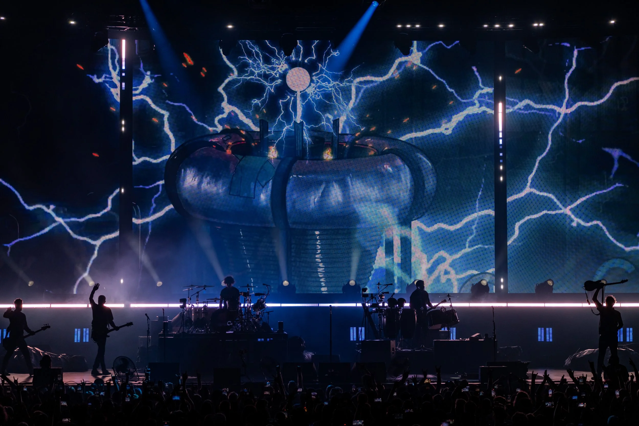 A concert stage with a band performing, silhouettes of band members, and a large LED screen displaying graphics of lightning and a giant mechanical heart with lightning bolts. Audience silhouettes at the bottom.