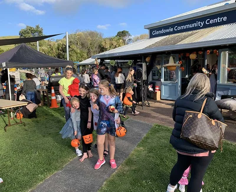 People gathered outside Glendowie Community Centre decorated for Halloween, with children holding pumpkin buckets, some dressed in costumes, and a tent set up on the grass.