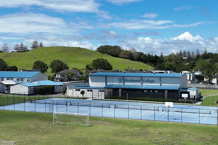 Outdoor sports facilities with tennis courts, a soccer goal, and a school building labeled 'Glendowie College' in the background, with green hills and trees under a blue sky with clouds.