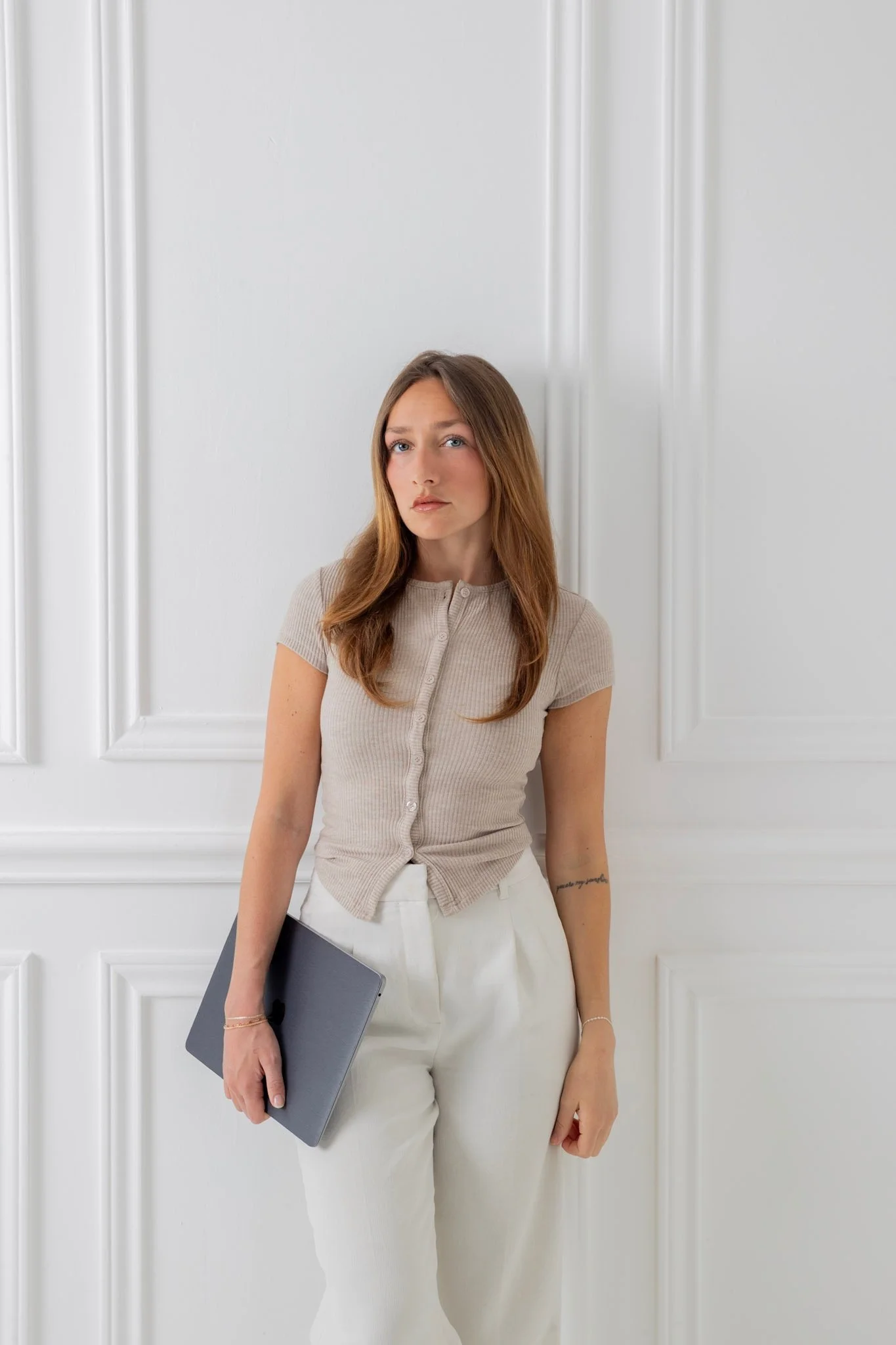 A young woman with long brown hair and blue eyes standing against a white paneled wall, holding a laptop in her right hand, wearing a beige short-sleeved button-up shirt and white high-waisted pants.