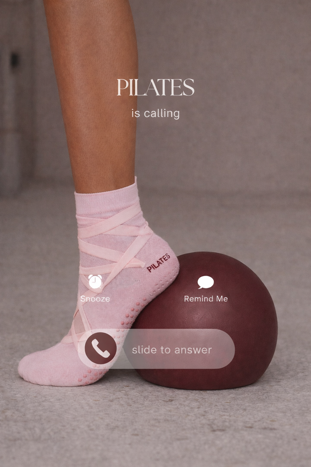 Close-up of a person's leg wearing a pink athletic sock with the word 'PILATES' on it, resting on a brown exercise ball, with a gray carpeted surface in the background.