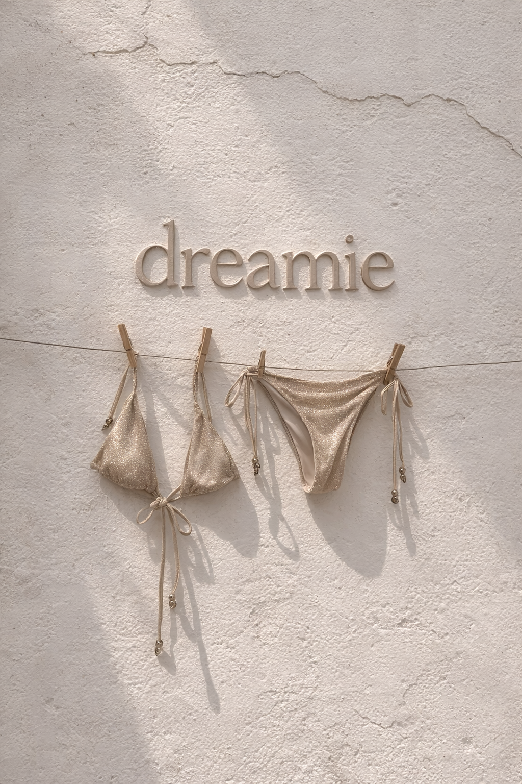 A beige, glittery bikini hanging on a string with clothespins, with the word 'dreamie' mounted on a textured white wall above it.