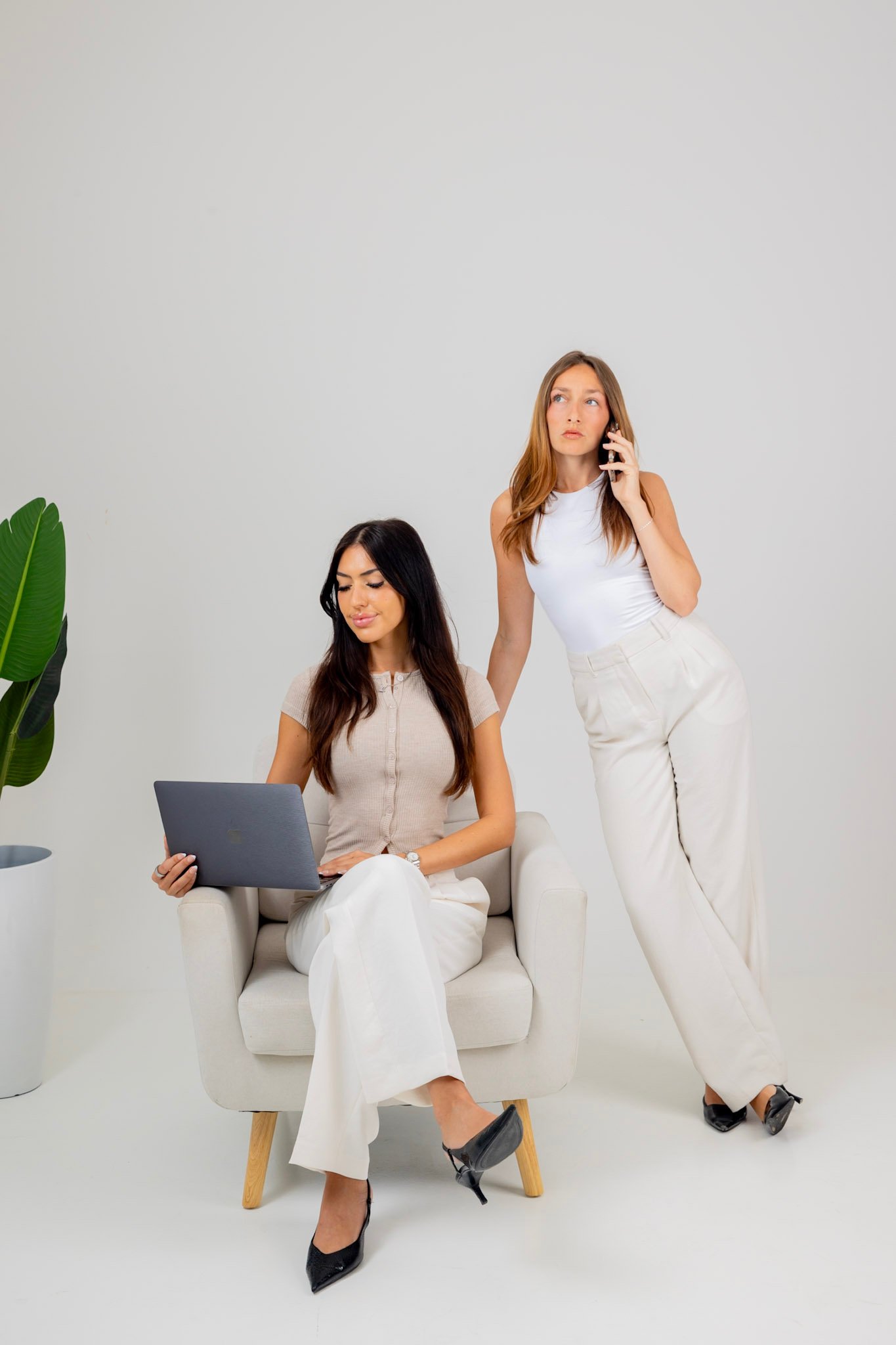 Two women, one seated with a laptop and the other standing, talking on a cellphone in a minimalistic room with a potted plant.
