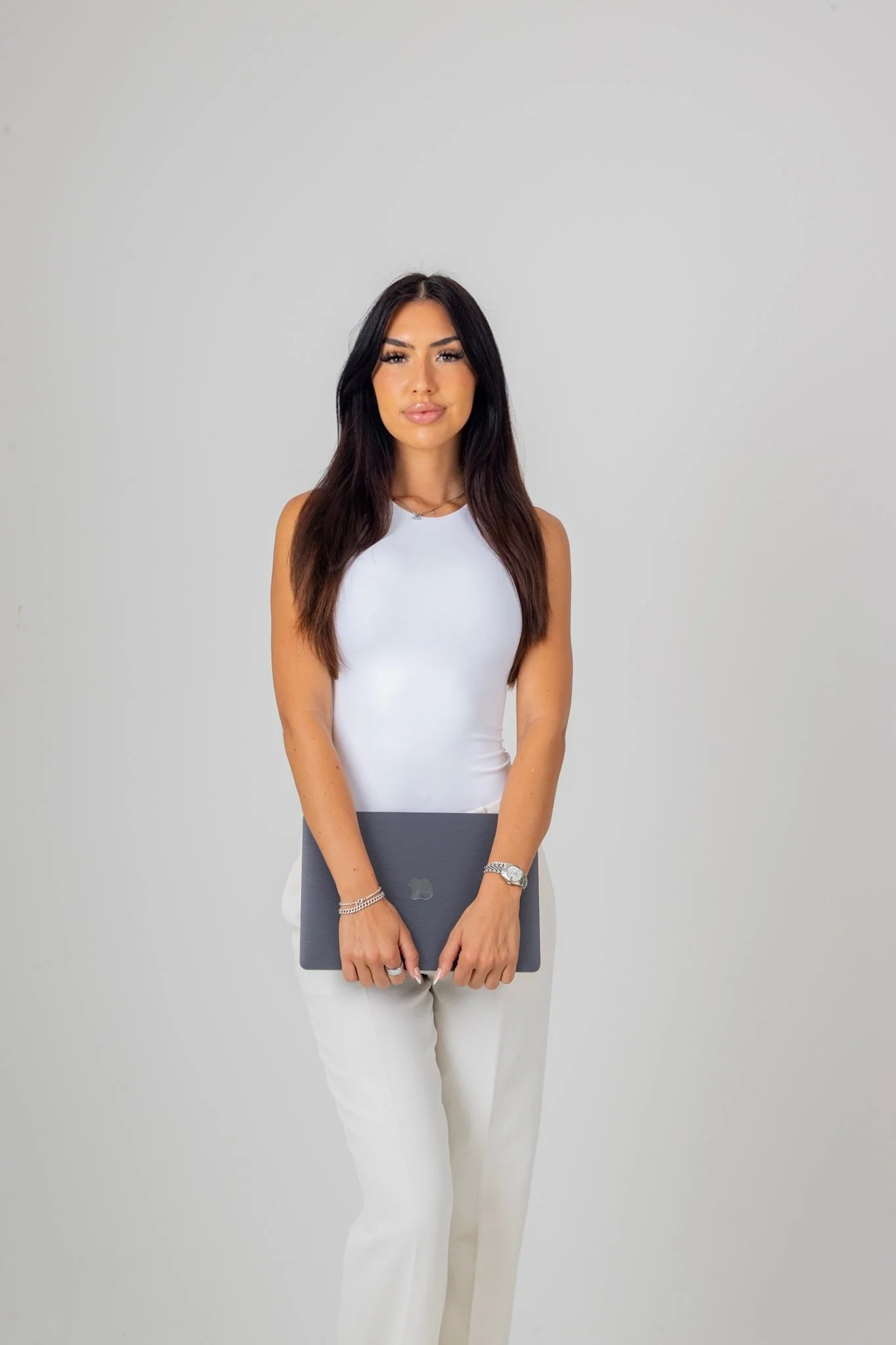 A woman with long dark hair and dressed in a white sleeveless top and white pants, holding a closed laptop in front of her, standing against a plain light gray background.
