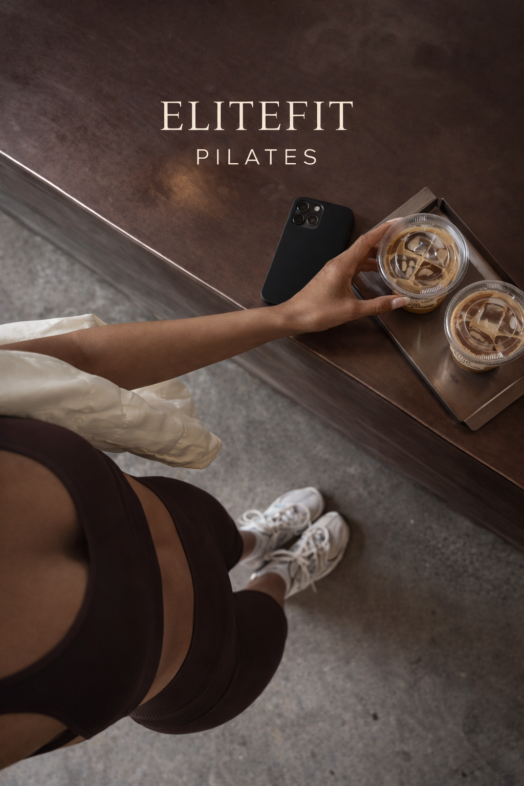 Top-down view of a woman standing in workout clothes, holding a plastic cup of iced coffee, with a smartphone on a wooden counter nearby. Two iced coffees are on a tray on the counter.