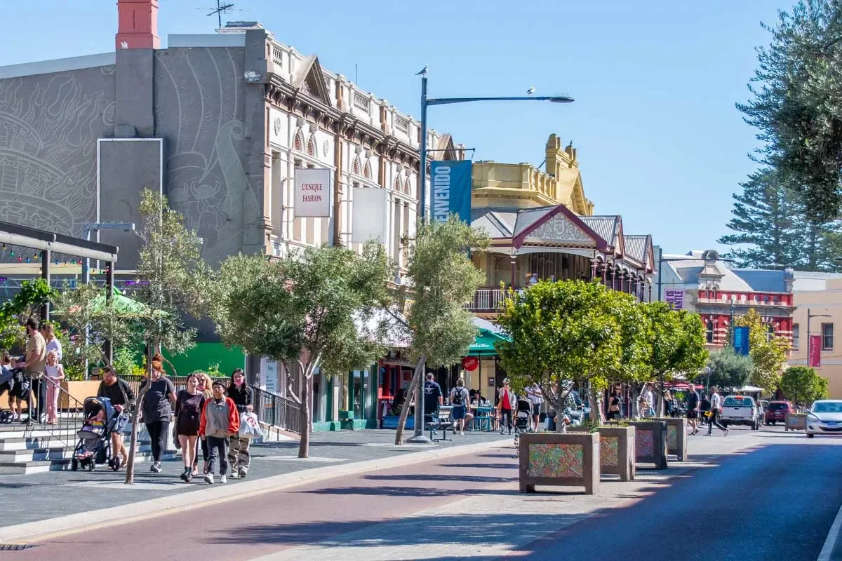 A lively street scene in a shopping district with people walking and shopping, storefronts, trees, and buildings in the background under a bright blue sky.