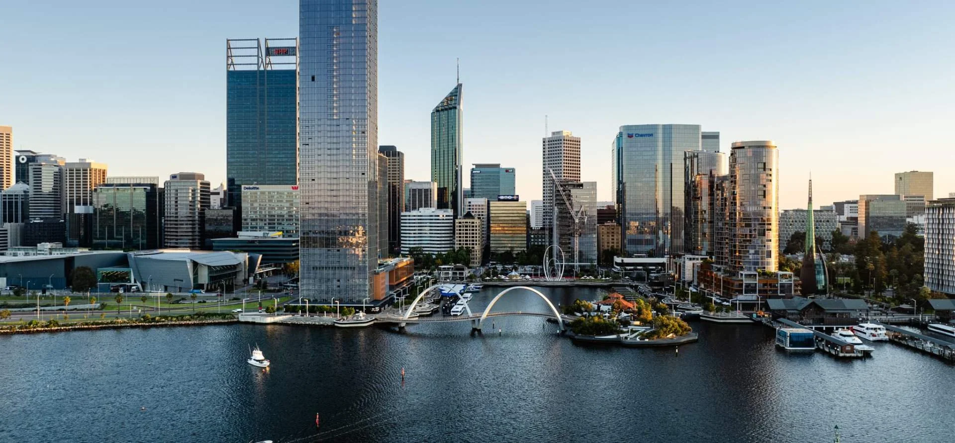 A city skyline with tall modern skyscrapers, a waterfront with boats, and an arched pedestrian bridge over the water, during daylight.