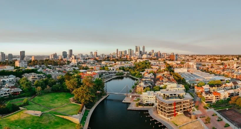 A city skyline with tall buildings in the distance, a river running through a lush green park with trees, and low-rise residential and commercial buildings in the foreground.