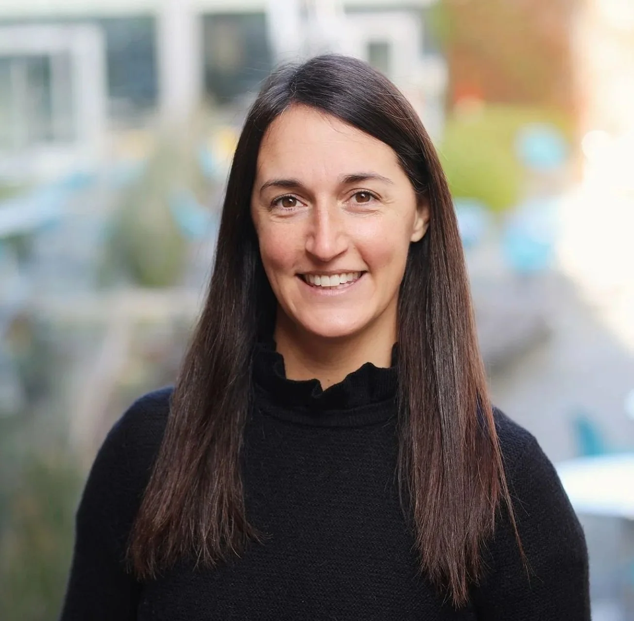 Professional woman with long brown hair smiling outdoors in front of a blurred background with buildings and trees.