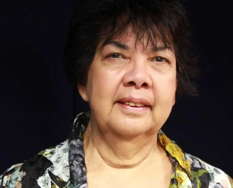 Close-up of an elderly woman with short dark hair, wearing a patterned shirt, against a dark background.
