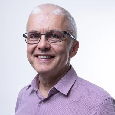 A smiling older man with glasses wearing a purple button-up shirt.