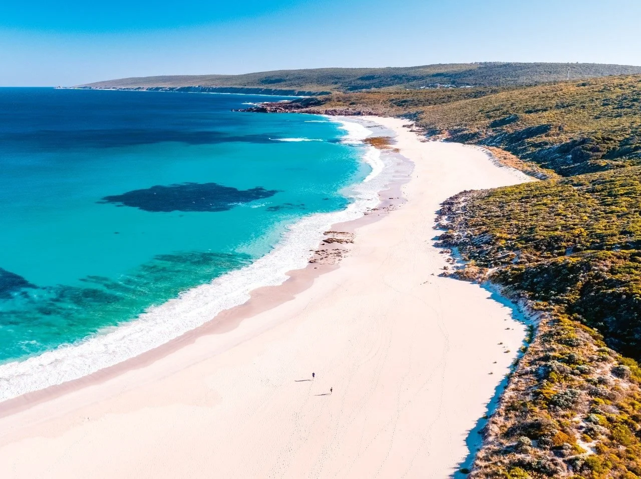 Aerial view of a long white sandy beach with clear turquoise ocean water and rocky green-covered hills in the background.