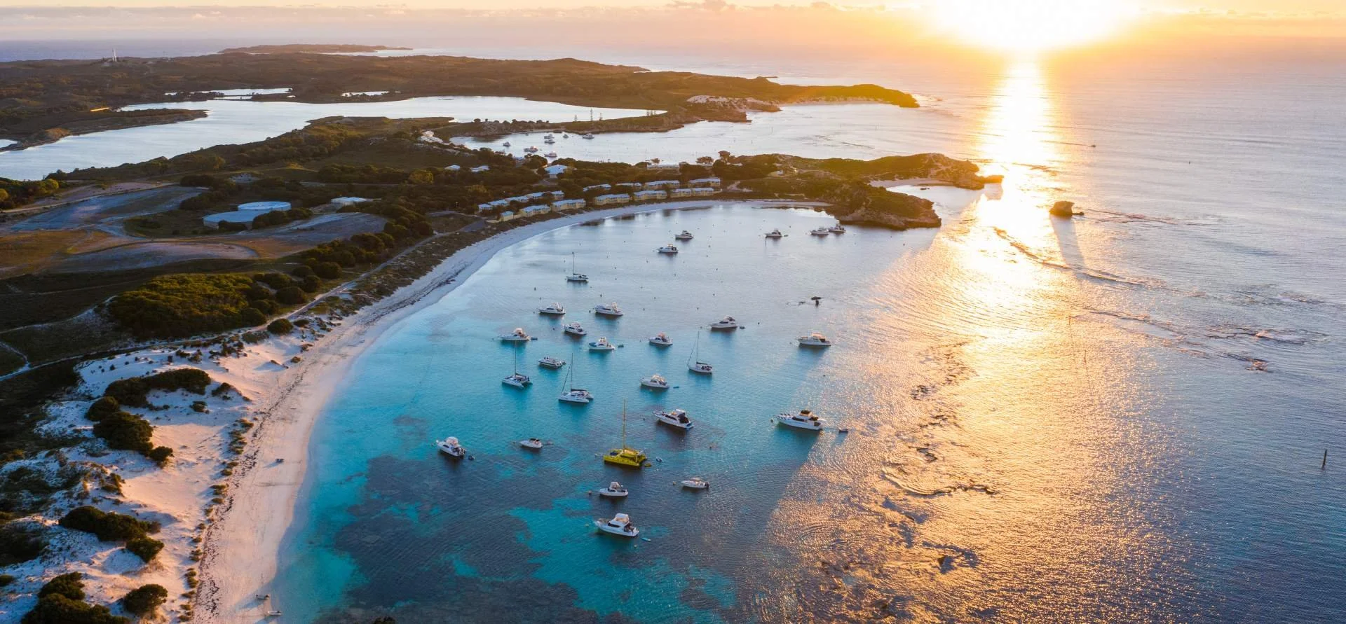 Aerial view of a coastal landscape at sunset with boats anchored in a cove and a sandy beach, surrounded by greenery and open water.