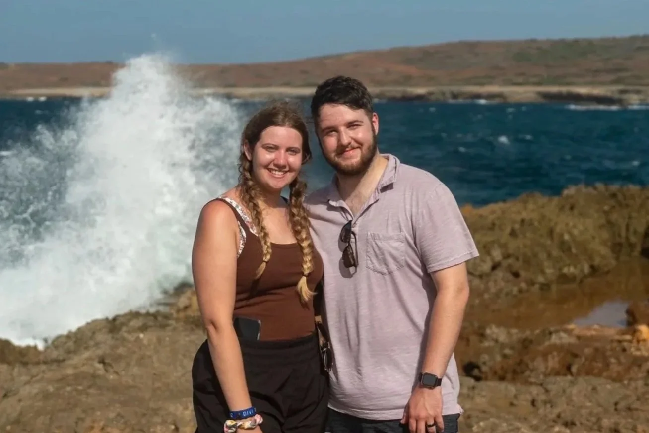 A young couple standing on rocky shoreline at the ocean, smiling, with waves crashing behind them and distant hills in the background.