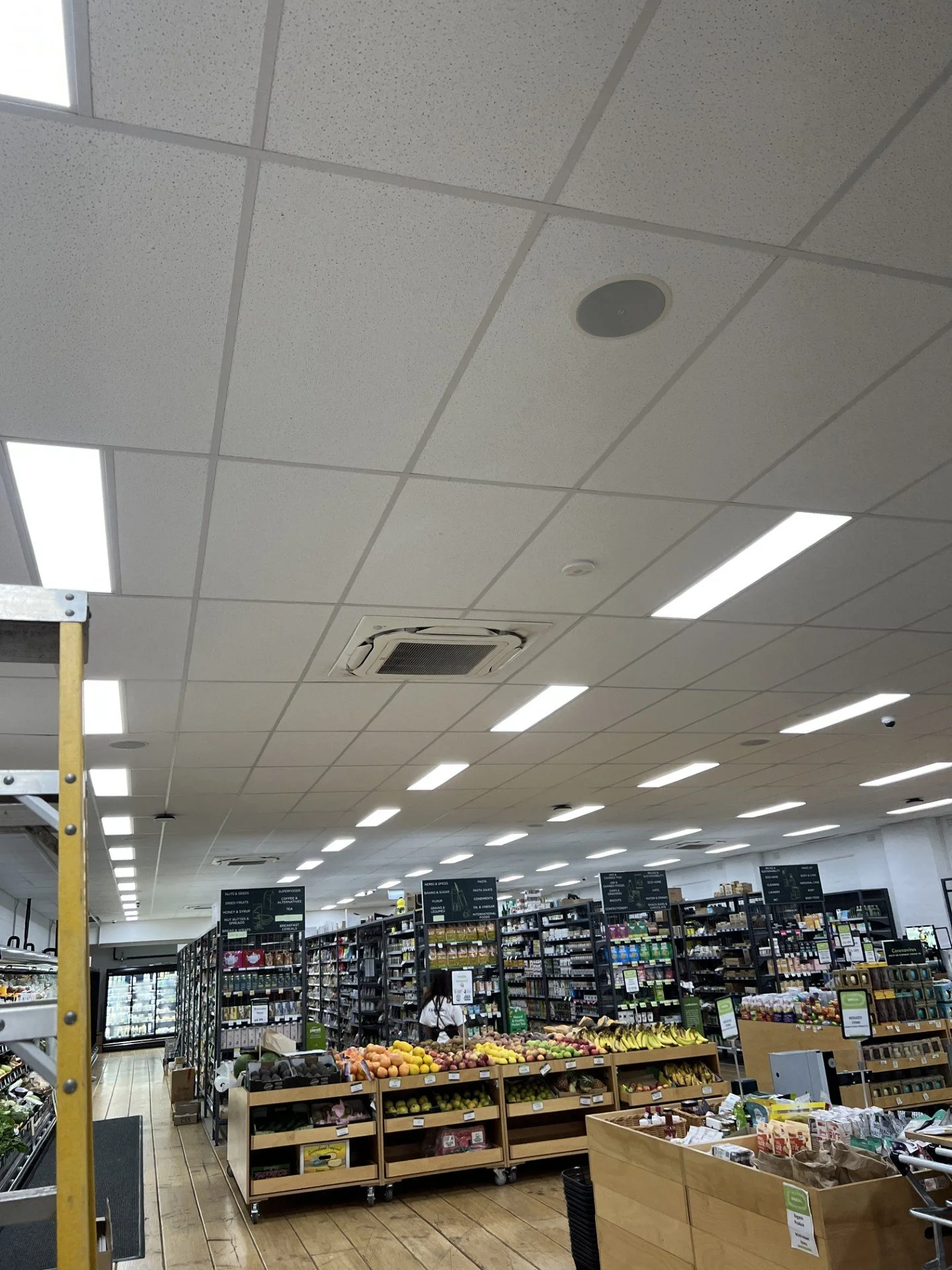 Interior of a grocery store with shelves of produce, snacks, and products, fluorescent lighting, and a ceiling with an air conditioning vent visible.