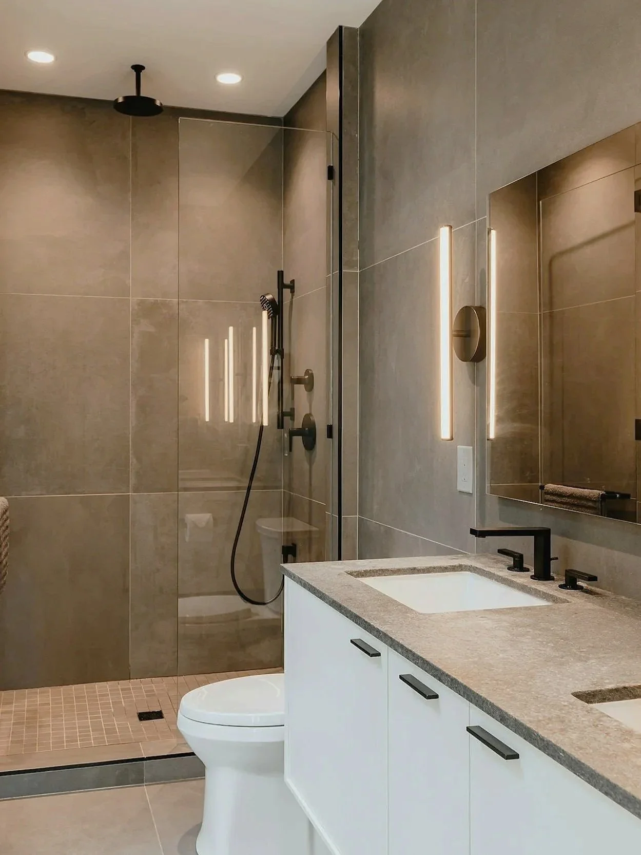 Modern bathroom with a walk-in shower featuring glass walls, a black rainfall showerhead, black fixtures, a white toilet, a vanity with a granite countertop, black faucets, and backlit mirror.