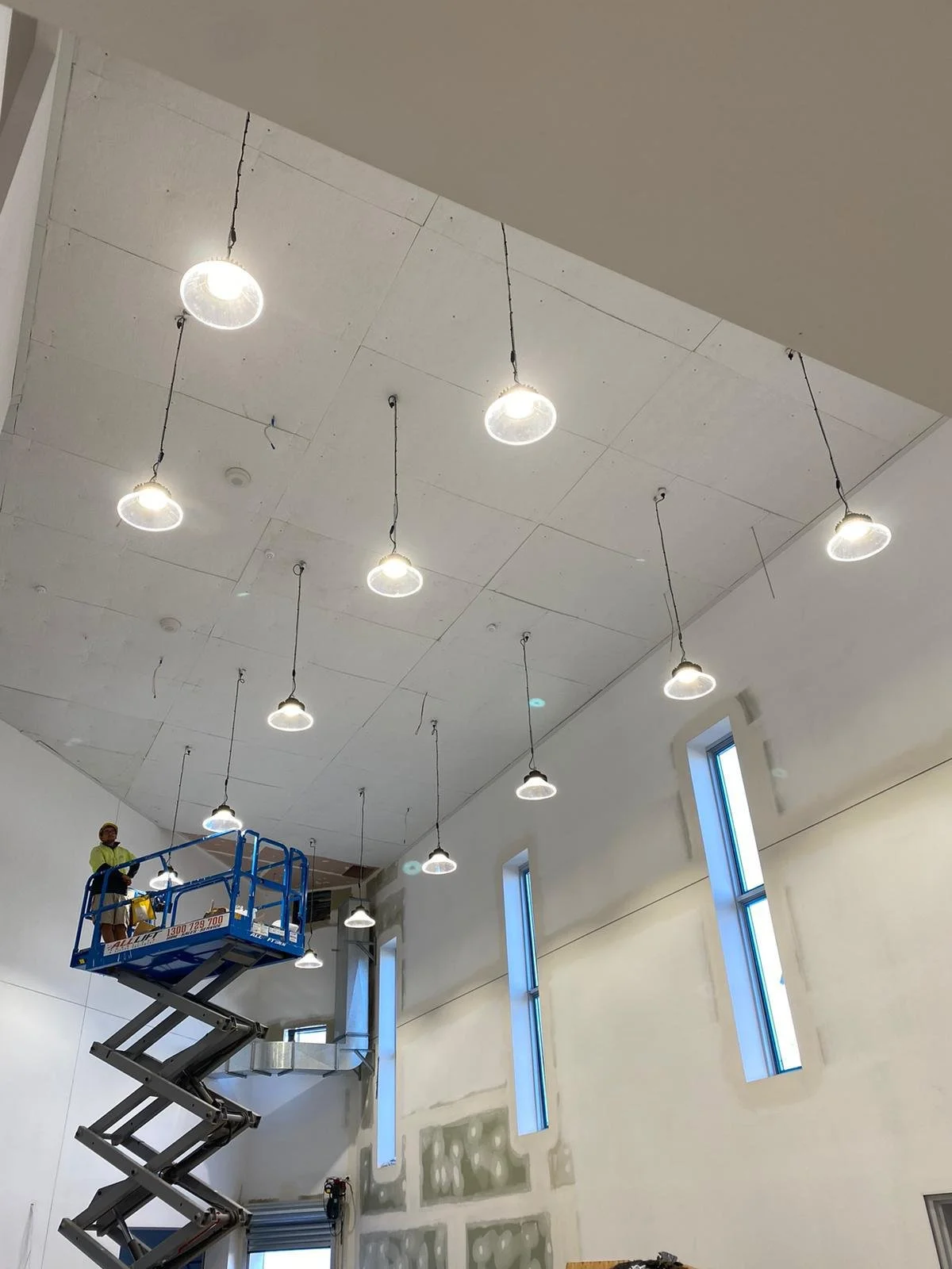 A worker on a blue scissor lift installing or maintaining ceiling lights in a large room with high windows and unfinished walls.