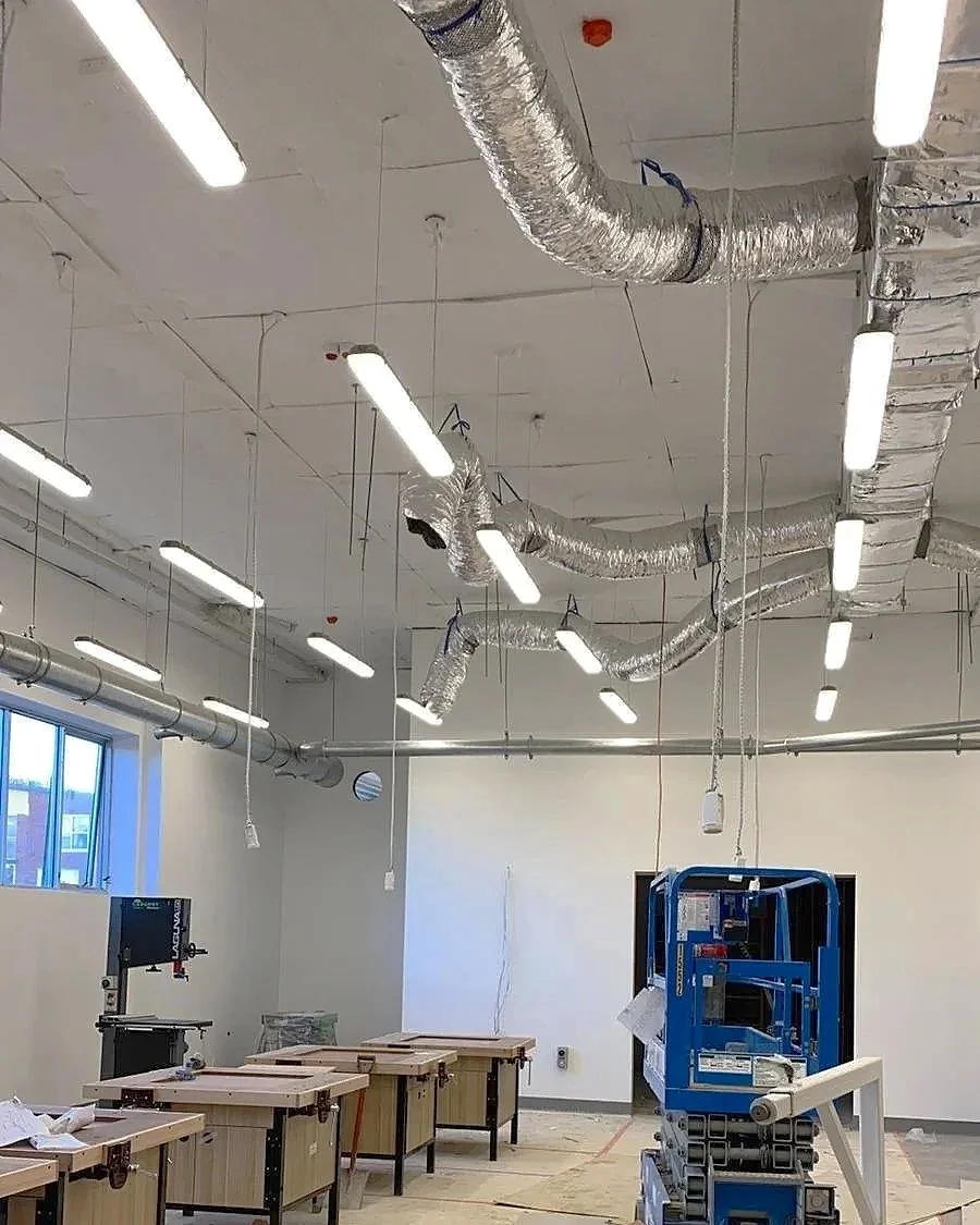 Construction or renovation site with exposed ductwork, hanging ceiling lights, and unfinished workstations or tables.