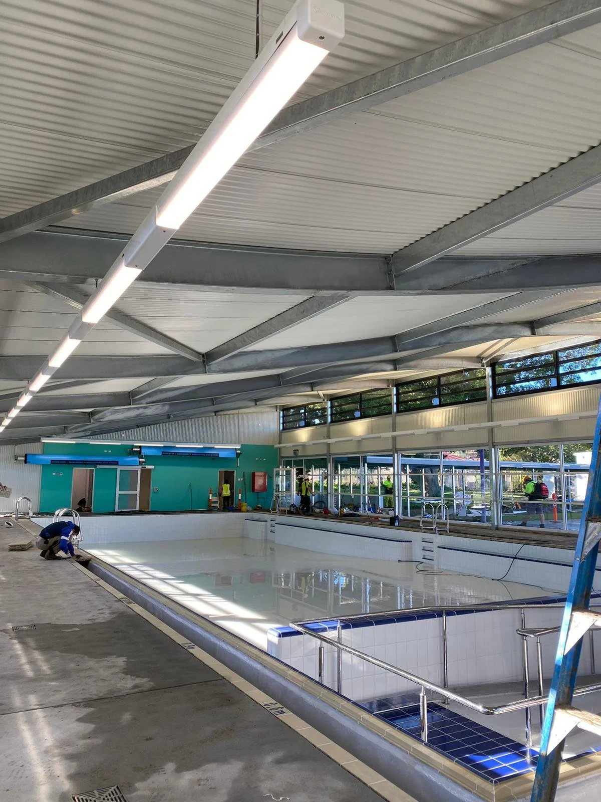 Indoor swimming pool under construction with workers installing and inspecting the area, surrounded by large windows and ceiling lights.