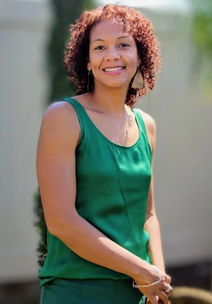 A woman with curly reddish-brown hair smiling outdoors, wearing a sleeveless green top and earrings.