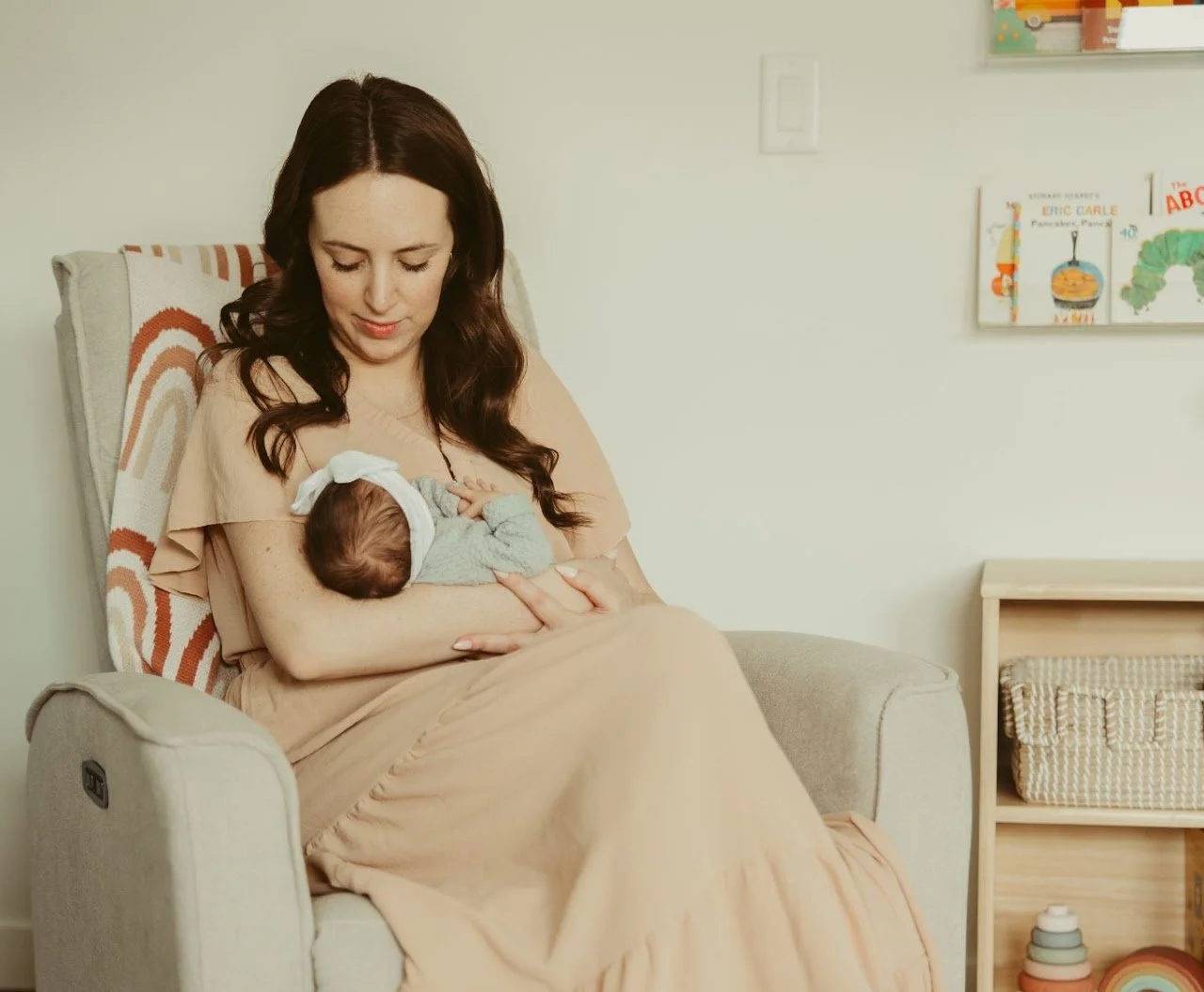 A woman with long dark hair sitting in a beige armchair breastfeeding a baby, who is wearing a gray outfit and a white headband. The background features a white wall with children's books and artwork.
