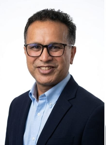 Portrait of a man with glasses wearing a dark blazer and light blue shirt, smiling against a white background.