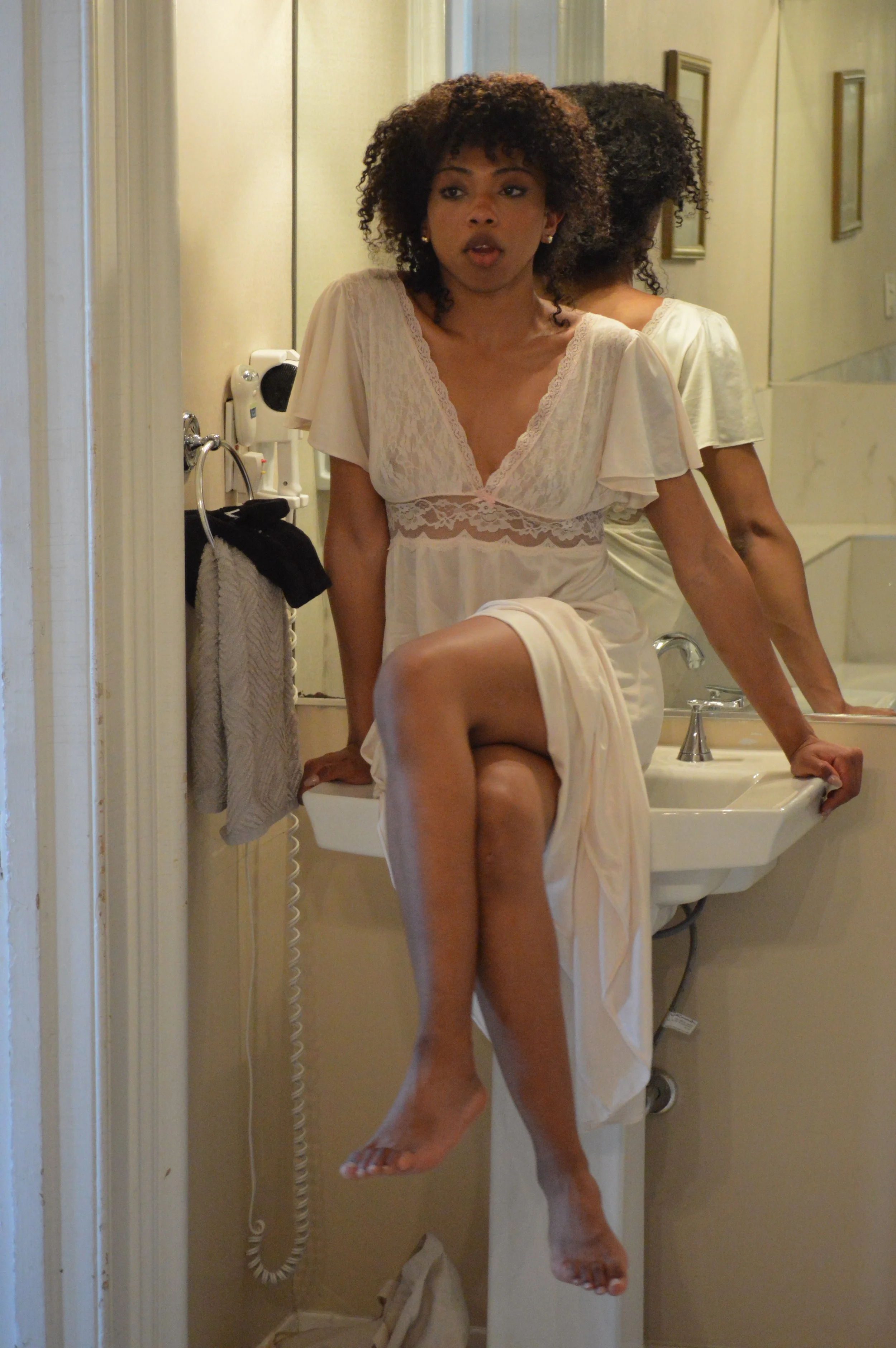 A woman with curly hair in a cream-colored nightgown sitting on a bathroom sink with one leg crossed. She is looking into a mirror, and her reflection is visible behind her. The bathroom has a mirror, a wall-mounted hairdryer, and a towel hanging on 