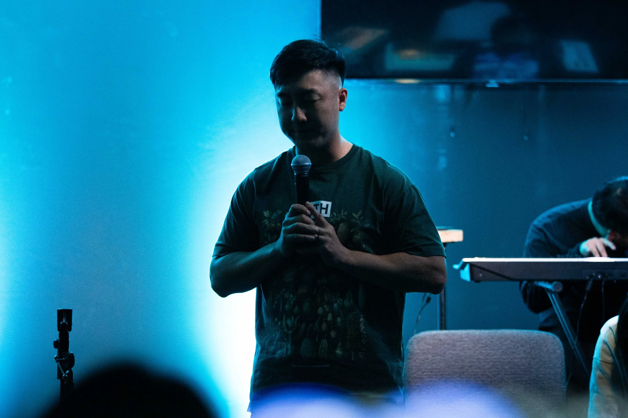 A young man with short dark hair, eyes closed, holding a microphone, standing in front of a blue-lit wall. In the background, there is a person playing a keyboard.