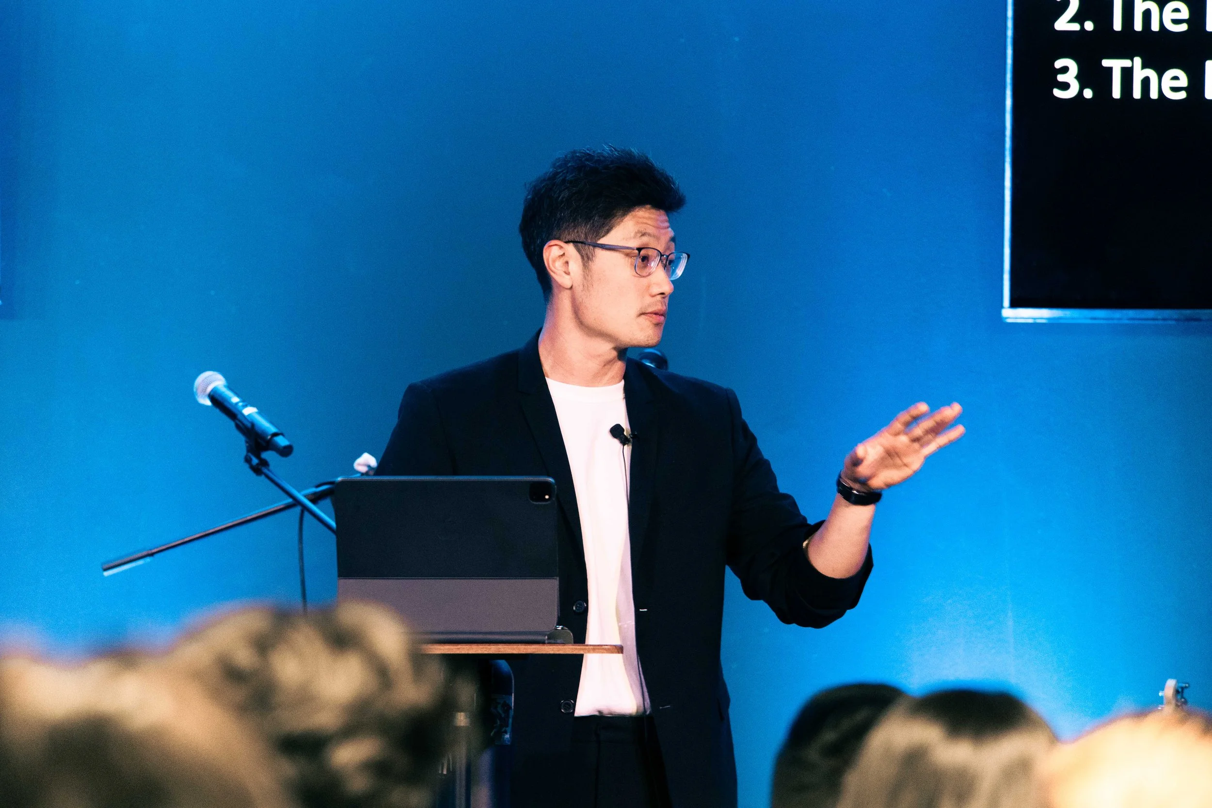 A man with black hair, glasses, wearing a black blazer and white shirt, is speaking at a podium during a presentation, with a blue background and part of a black screen with white text visible.