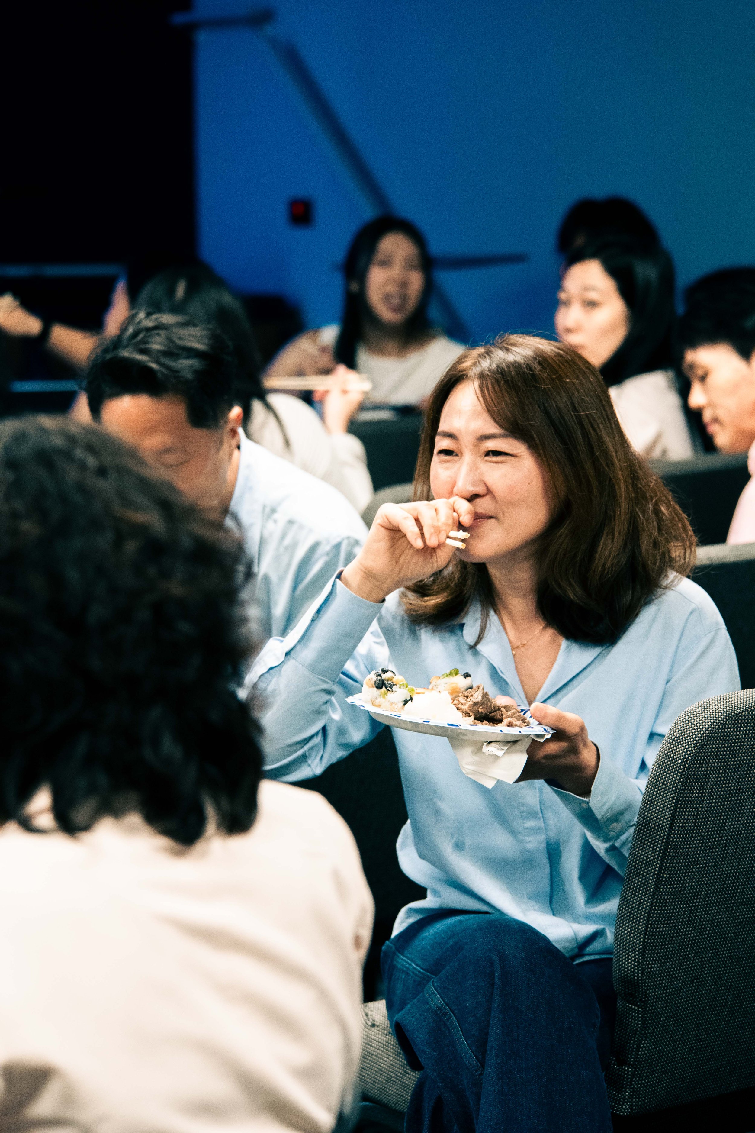A woman with shoulder-length brown hair wearing a light blue shirt holding a plate of food, smiling, in a busy indoor setting with other people.