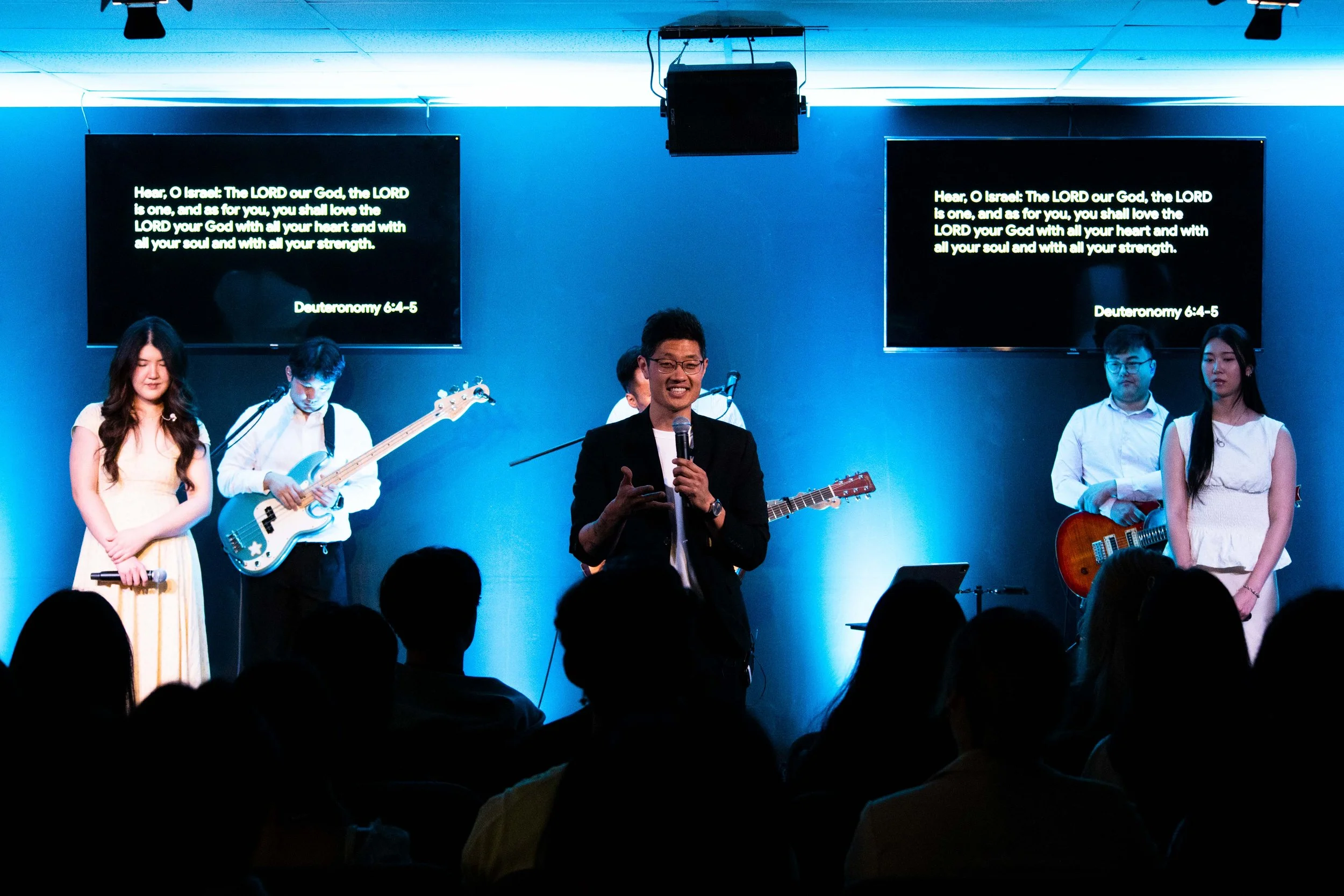 A group of musicians and a speaker on stage during a worship service, with two screens displaying biblical text.