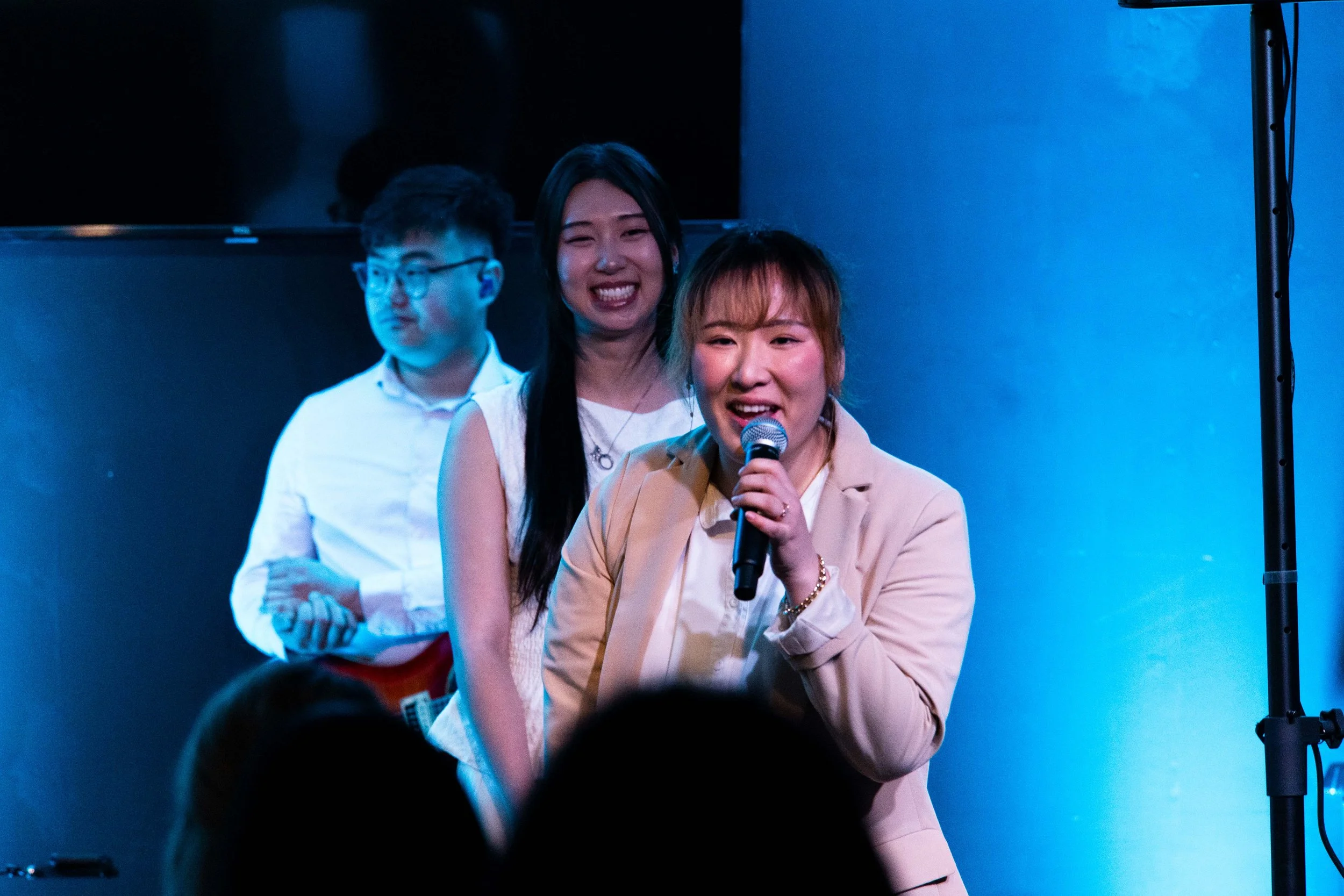 Three people on stage, a woman speaking into a microphone, smiling, with two young women standing behind her. The background is blue and black.