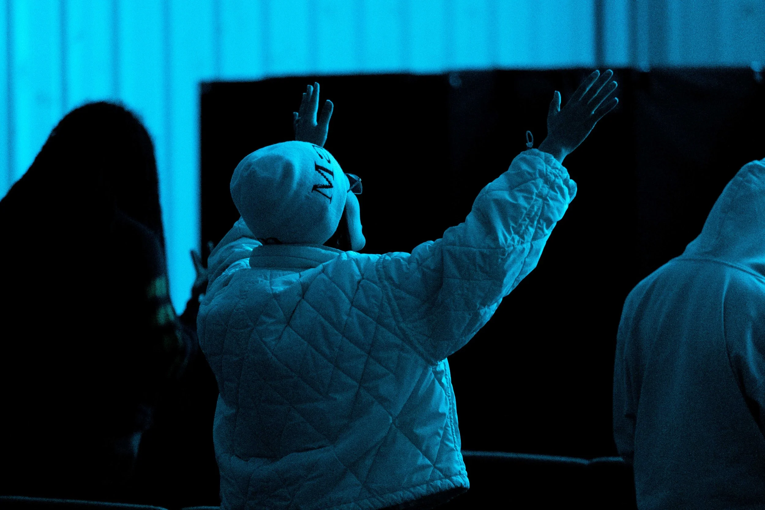 Person wearing a white quilted jacket and a white beanie with the word 'WAVE' on it is raising their hand, facing away from the camera, in a dimly lit room with blue lighting.
