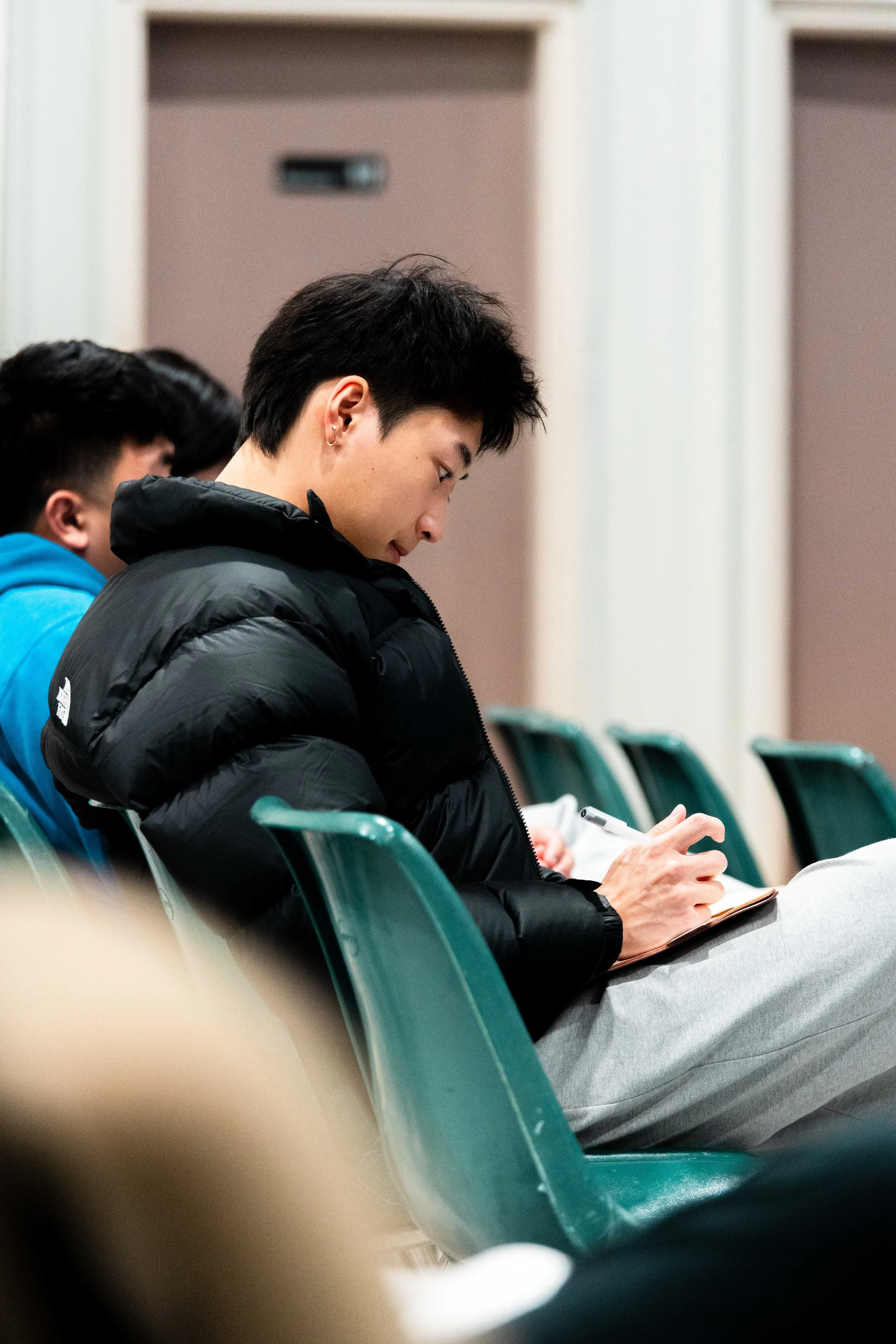 Side view of a young man with black hair, earrings, wearing a black jacket, sitting in a row of teal chairs, writing or drawing in a notebook with a pen.