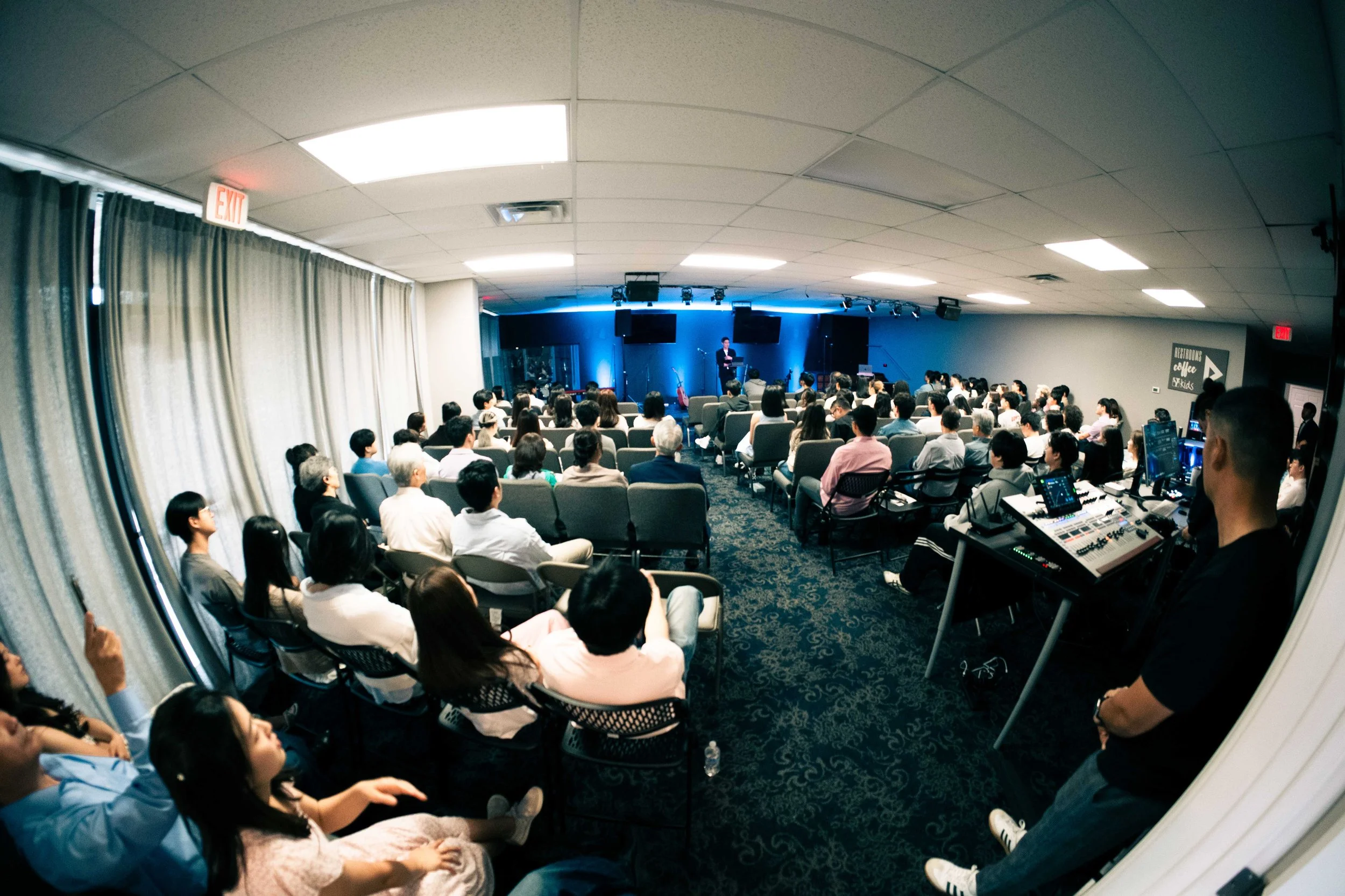 An audience sitting in chairs watching a speaker on stage at a conference or presentation, with a band setup behind the speaker and a blue-lit backdrop.
