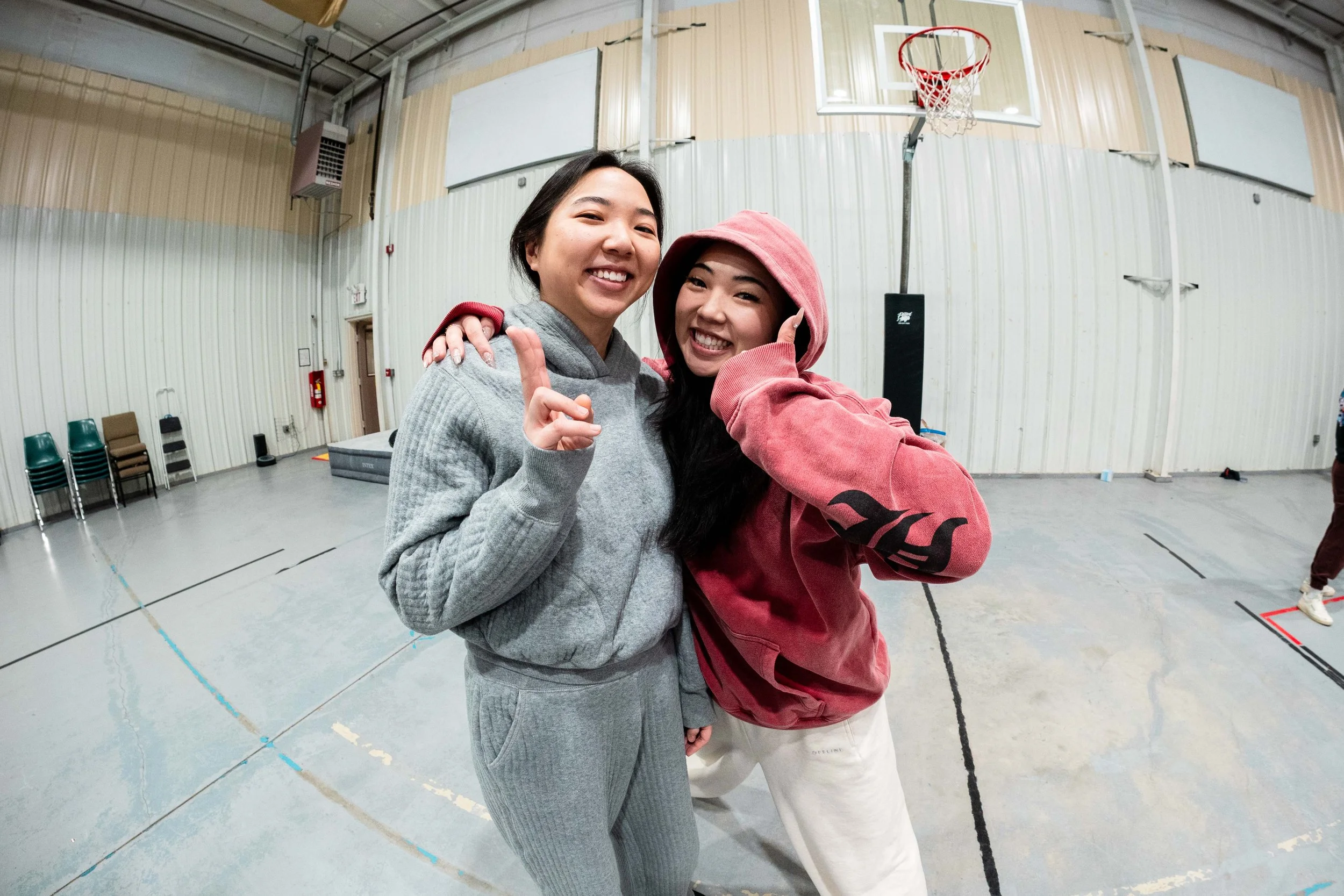 Two women smiling and posing inside a gymnasium near a basketball hoop, wearing casual sportswear.