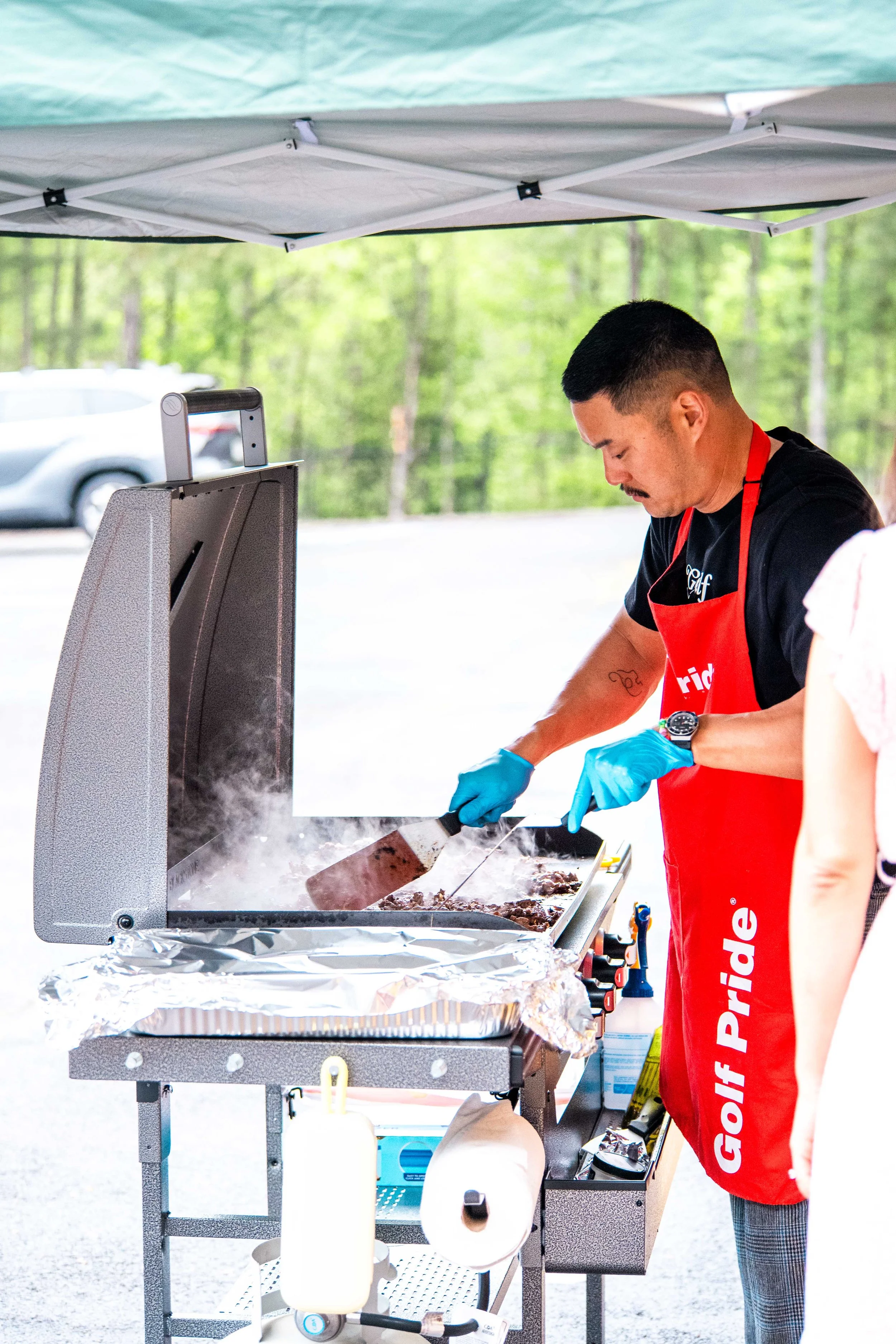 Man wearing a red apron and blue gloves grilling meat on a stainless steel barbecue grill outdoors, with green trees in the background and a white canopy overhead.