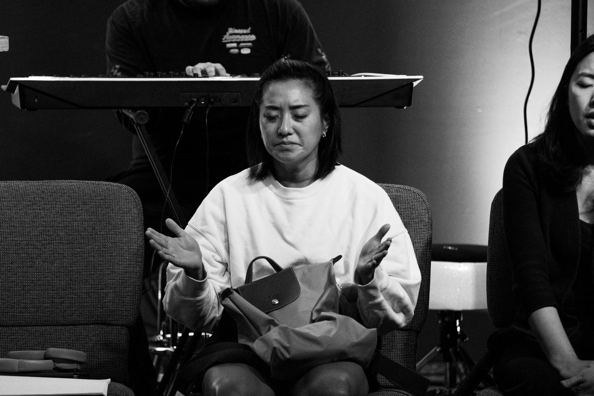 A black and white photo of a woman sitting with her eyes closed and arms crossed, with a man behind her using a keyboard.