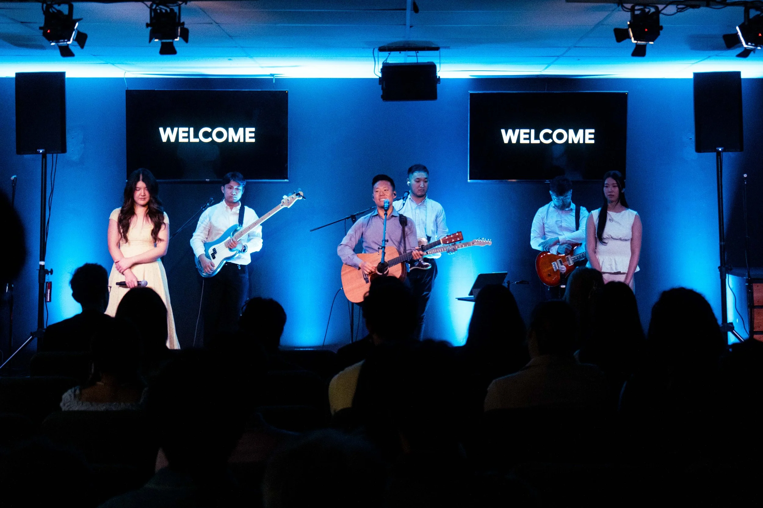 A group of six young musicians performing on a stage with blue and black background. Two women and four men, some holding guitars and one with a bass guitar. The stage has two large screens displaying the word 'WELCOME'. Audience silhouettes are visi