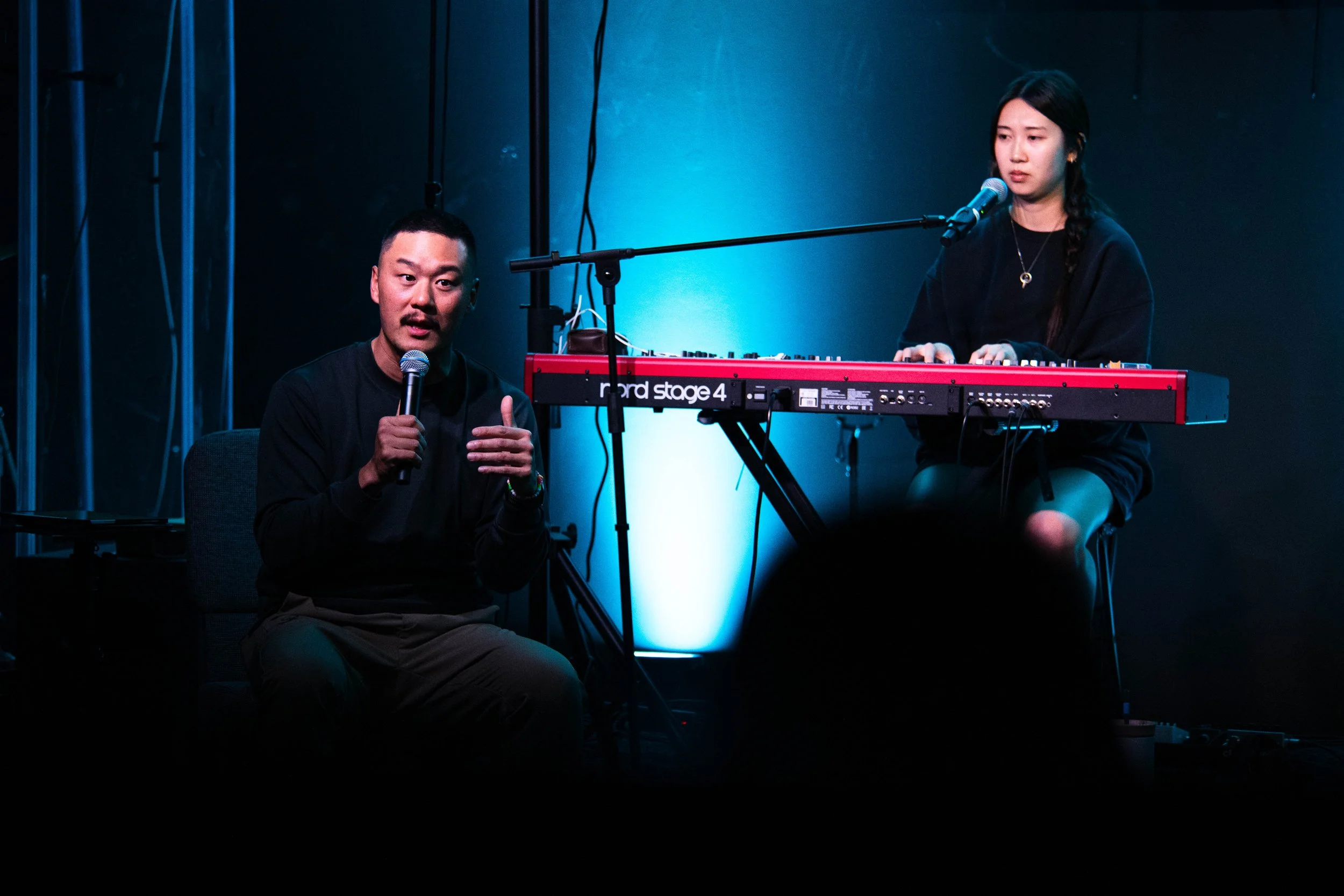 A man and a woman are performing music on stage. The man is sitting and speaking into a microphone, while the woman is playing a keyboard and singing into a microphone. The stage is lit with blue lighting.