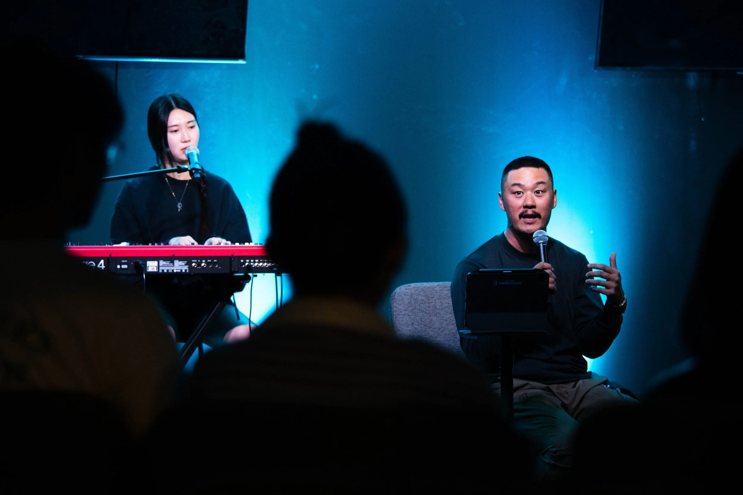 A woman playing keyboard and singing into a microphone, and a man speaking into a microphone while seated on stage in a dark room with blue lighting.