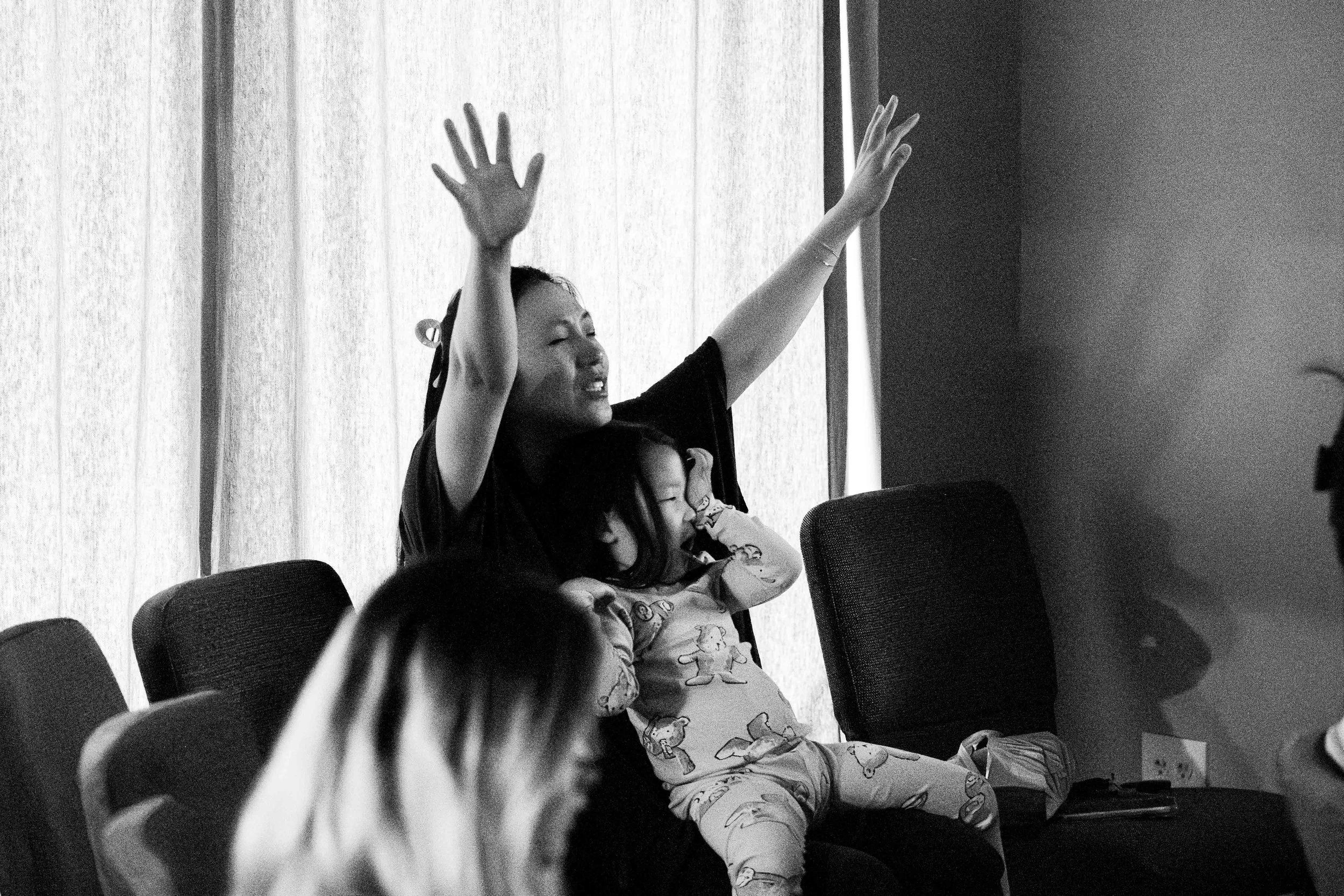 A woman with eyes closed and arms raised, sitting in a chair with children around her, in a room with curtains and chairs, captured in black and white.